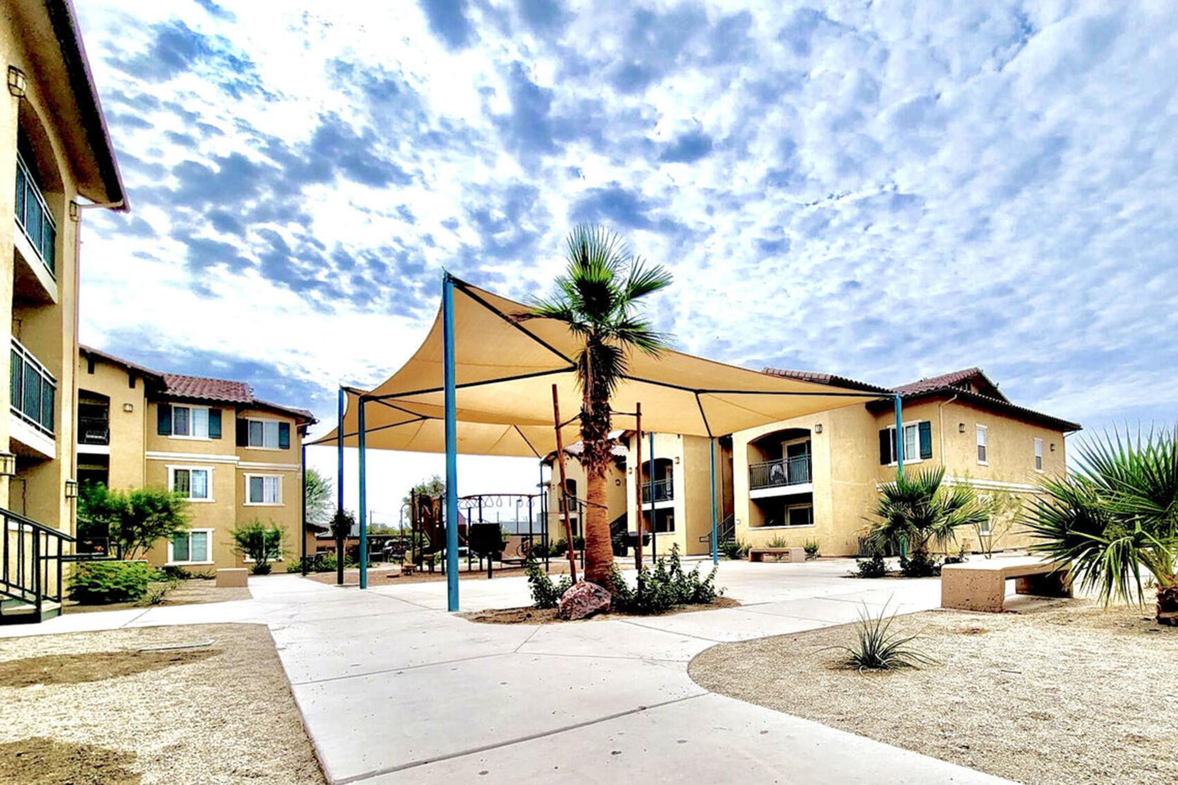A landscaped courtyard featuring a large shaded structure under a cloudy sky, surrounded by residential buildings. The area includes palm trees, shrubs, and a pathway, creating a relaxing outdoor space in a residential complex.