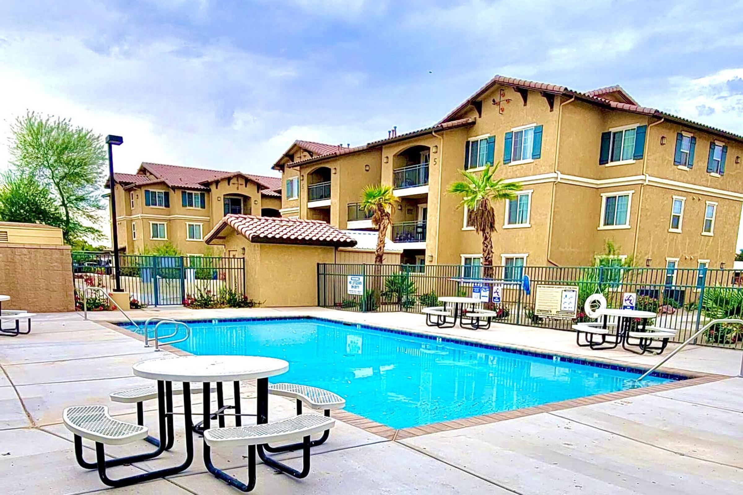 A clean and inviting pool area with several round tables and chairs surrounding the pool. In the background, a multi-story residential building features palm trees and a well-maintained courtyard. The sky is partly cloudy, creating a pleasant atmosphere.