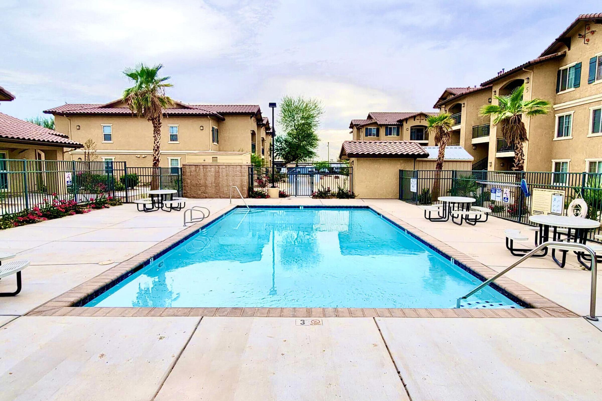 A clear swimming pool surrounded by beige apartment buildings. The pool area features several tables and chairs along the sides, along with palm trees and vibrant landscaping. The sky is overcast, adding a serene atmosphere to the scene.