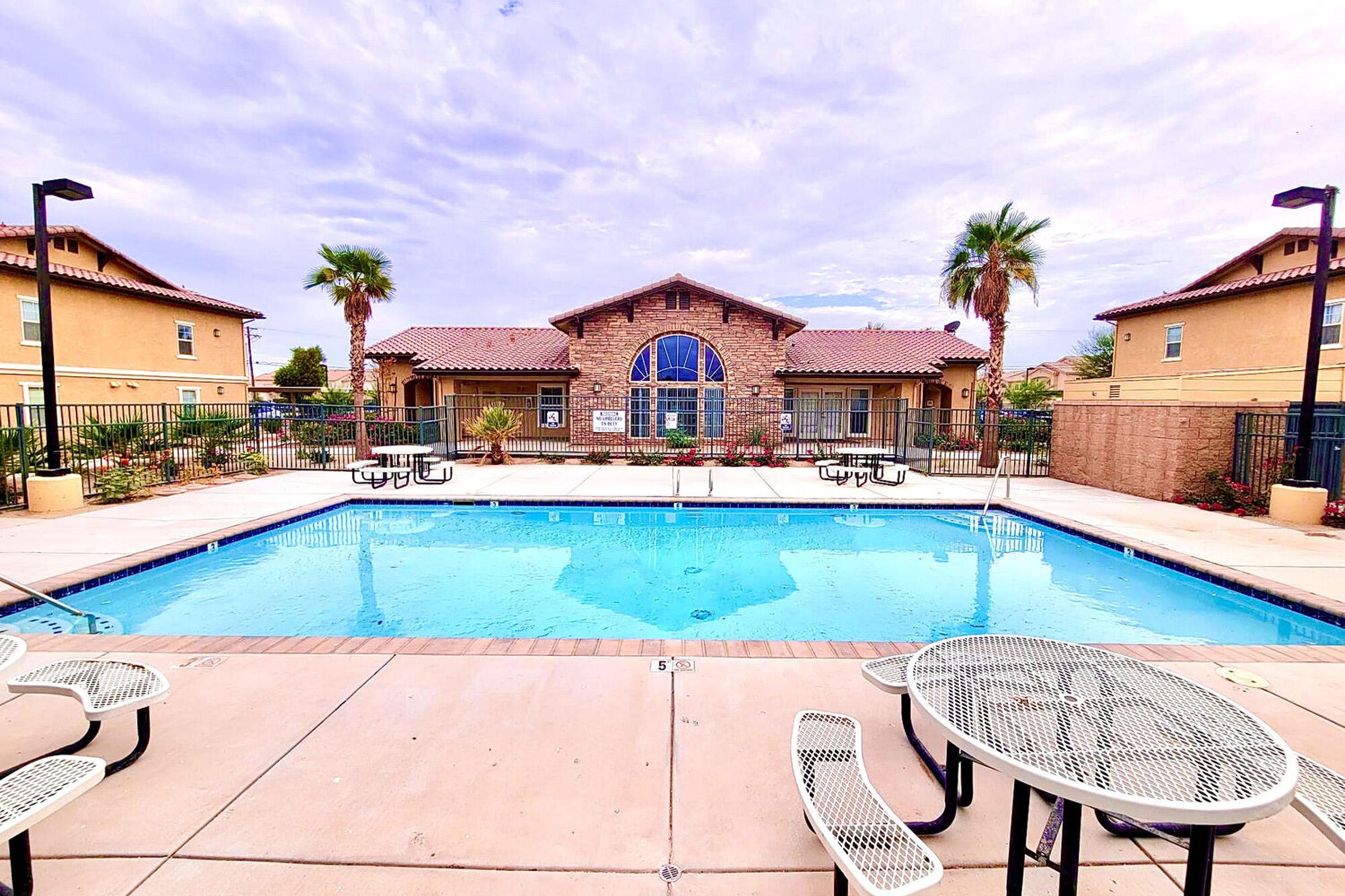A well-maintained outdoor pool surrounded by lounge chairs, tables, and palm trees, with a clubhouse in the background. The sky is partly cloudy, and the area appears inviting and spacious, ideal for relaxation and leisure activities.