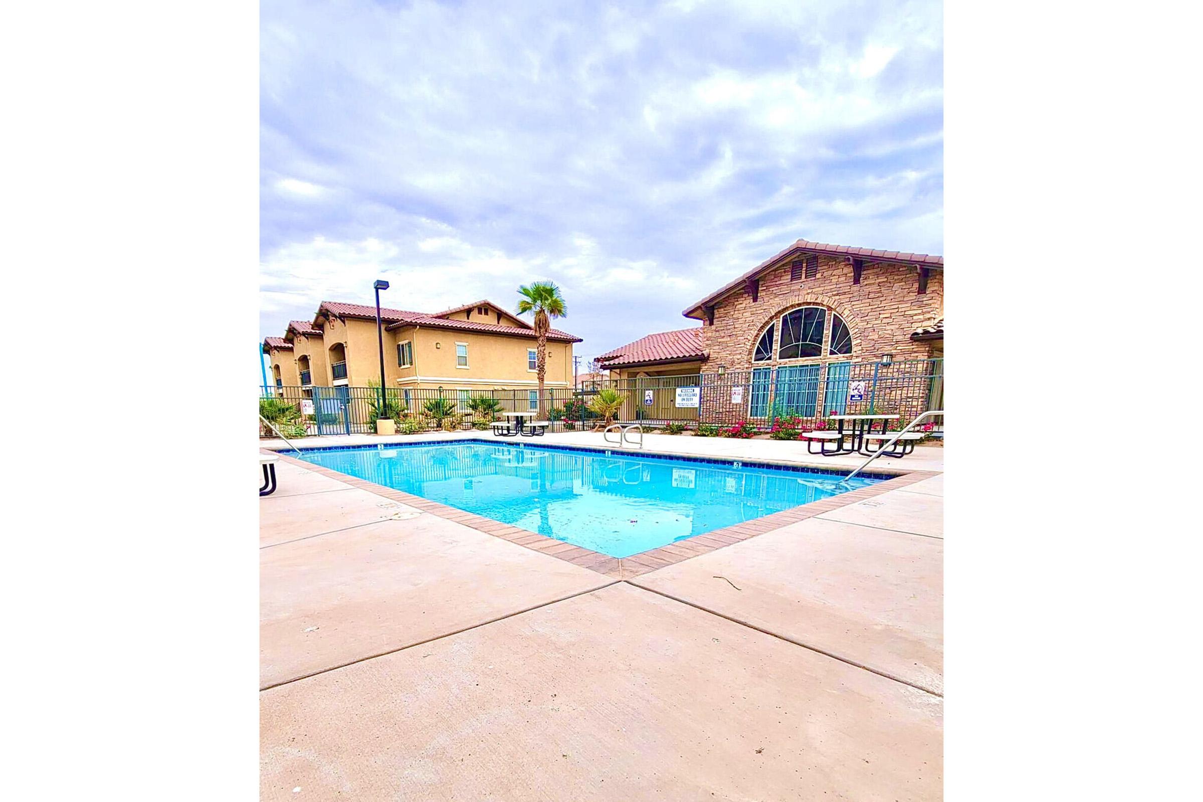 A clear pool surrounded by a concrete deck, with lounge chairs on either side. In the background, there are residential buildings with a mix of stone and stucco exteriors, and landscaping with palm trees and flowers. The sky is partly cloudy, hinting at a warm day.