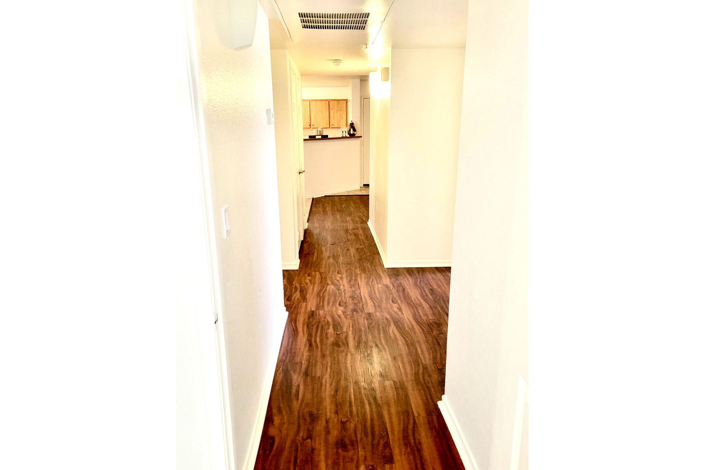 A brightly lit interior hallway featuring light-colored walls and wooden flooring. The corridor leads towards an open kitchen area visible in the distance, with a smooth curve on the right side. The space appears clean and modern, creating an inviting atmosphere.