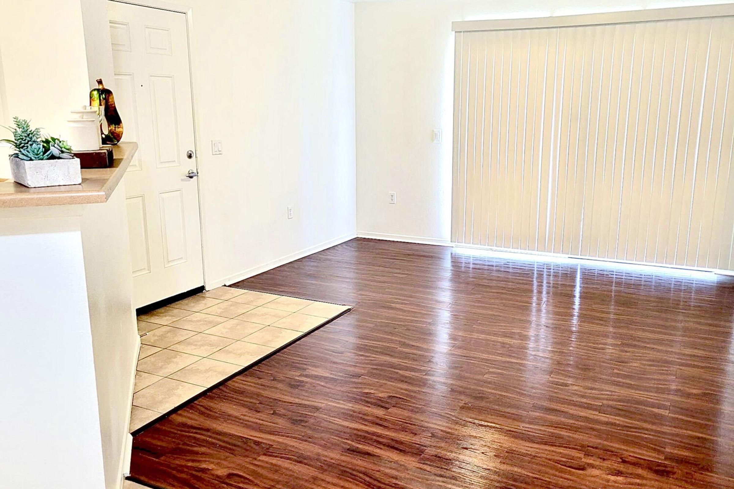A spacious, well-lit interior of a room featuring glossy wooden flooring, a doorway on the left, and a large window with vertical blinds allowing natural light. A small plant sits on a counter, adding a touch of decor to the otherwise empty space.