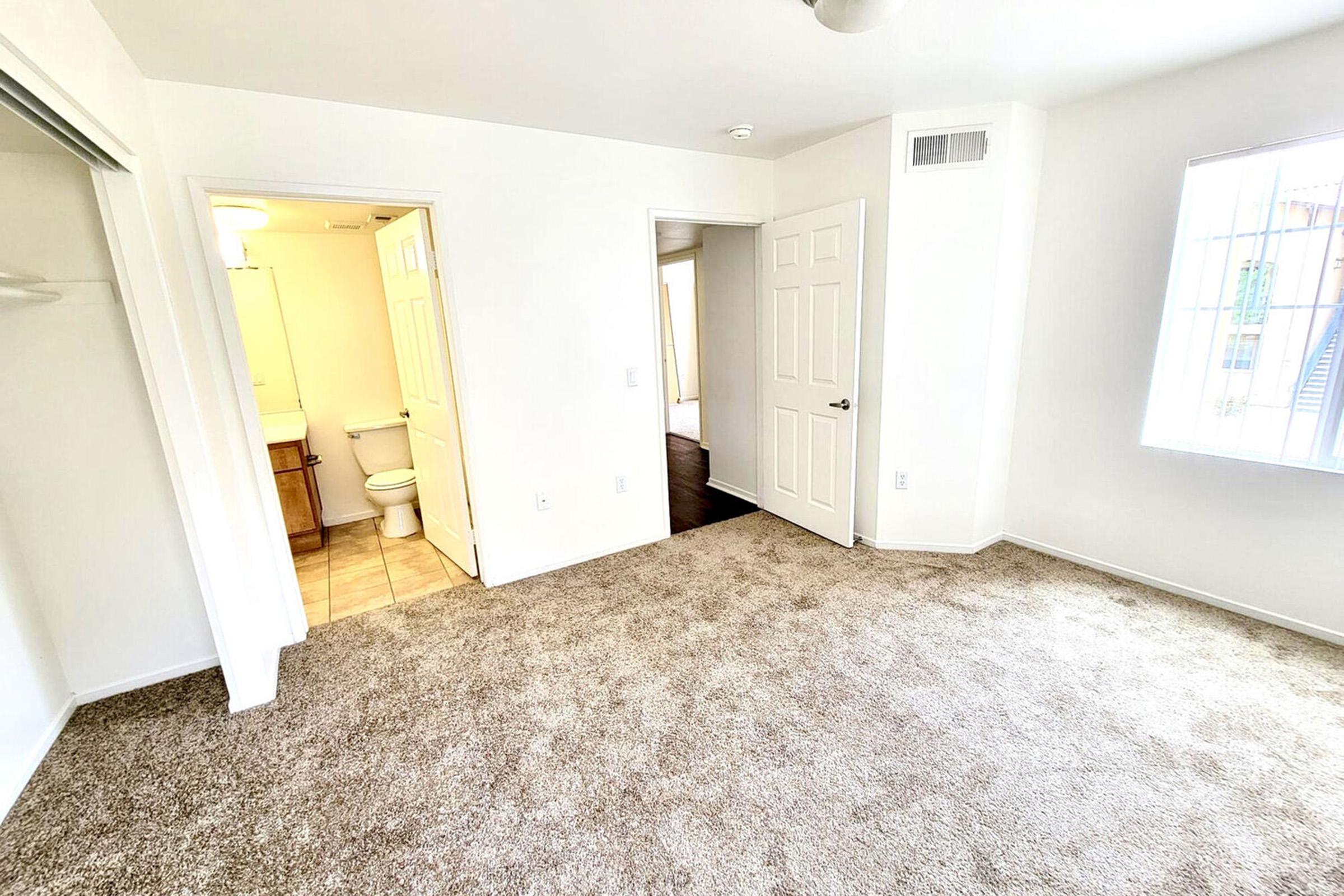 A bright, empty room with beige carpeting. Two doorways lead to a bathroom and another room. A window on the far wall allows natural light, and there is a ceiling fan above. The walls are painted white, creating a clean and spacious feel.