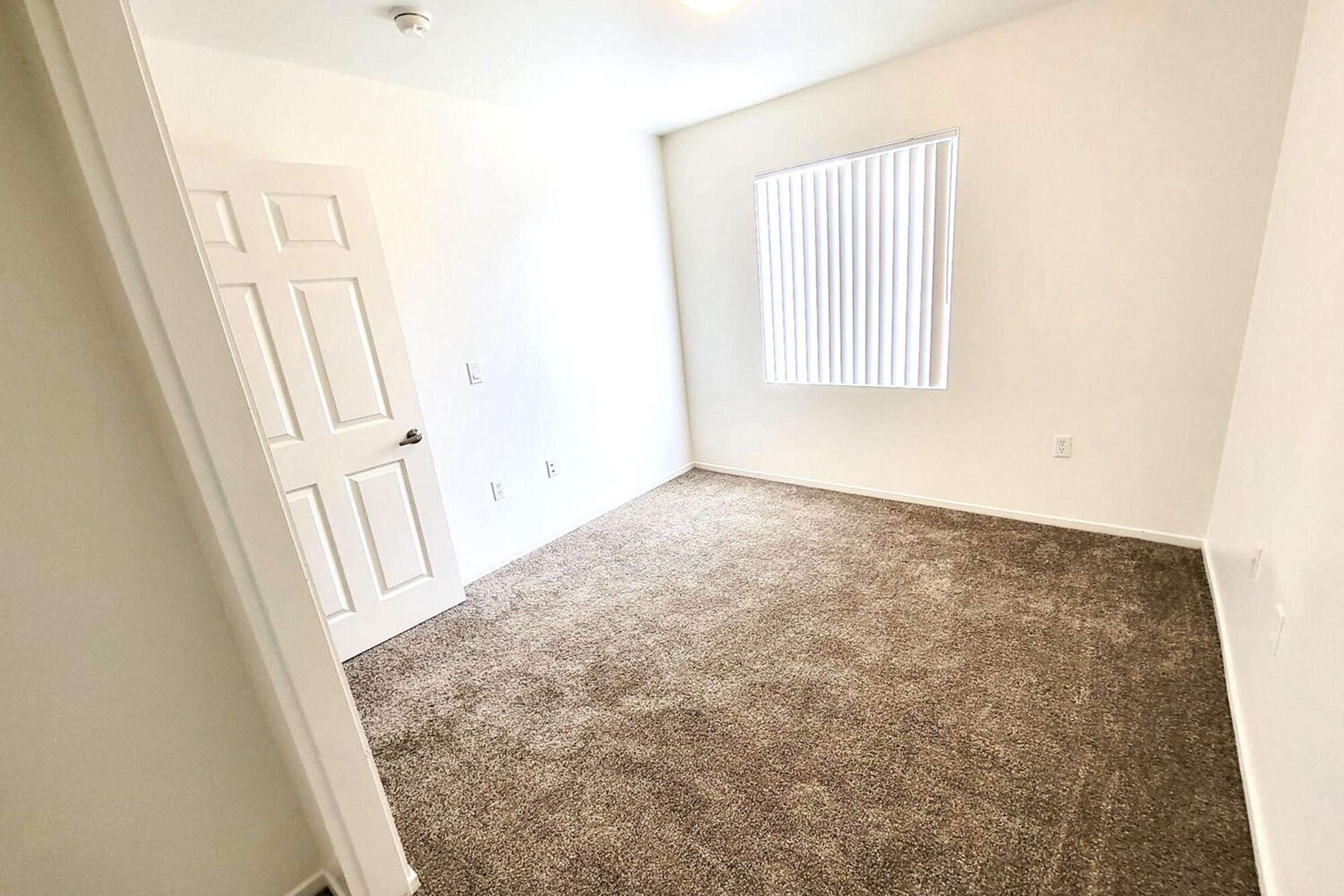 Empty room with light-colored walls and a carpeted floor. A single window with vertical blinds allows natural light to enter. A door is visible on the left side of the image, leading to another area. The space is clean and uncluttered, suggesting it may be used for various purposes.