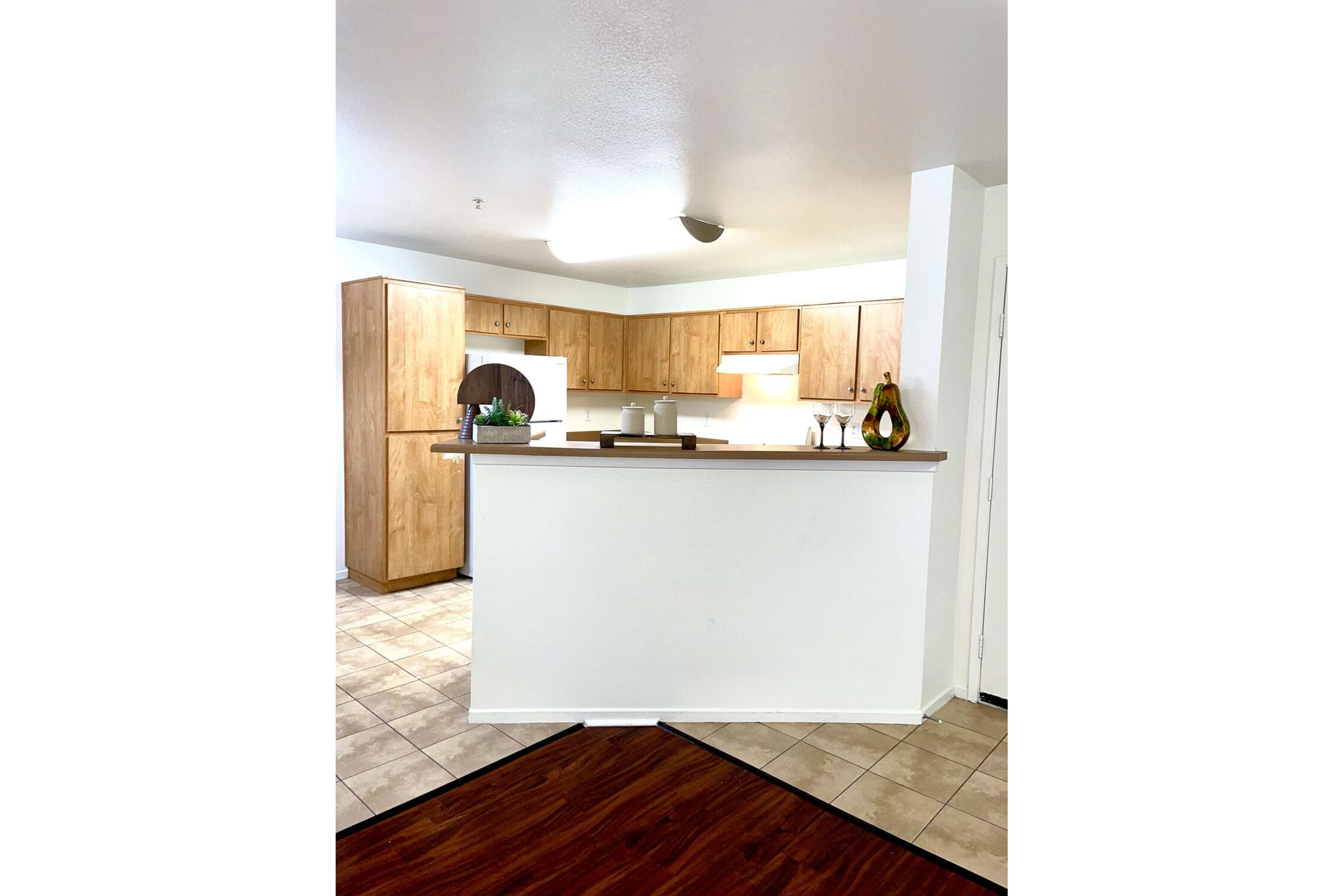 A modern kitchen with wooden cabinetry, a countertop that separates the kitchen from a dining area, and tile flooring. The space features a small breakfast bar with decorative items and ambient lighting, creating a warm and inviting atmosphere.