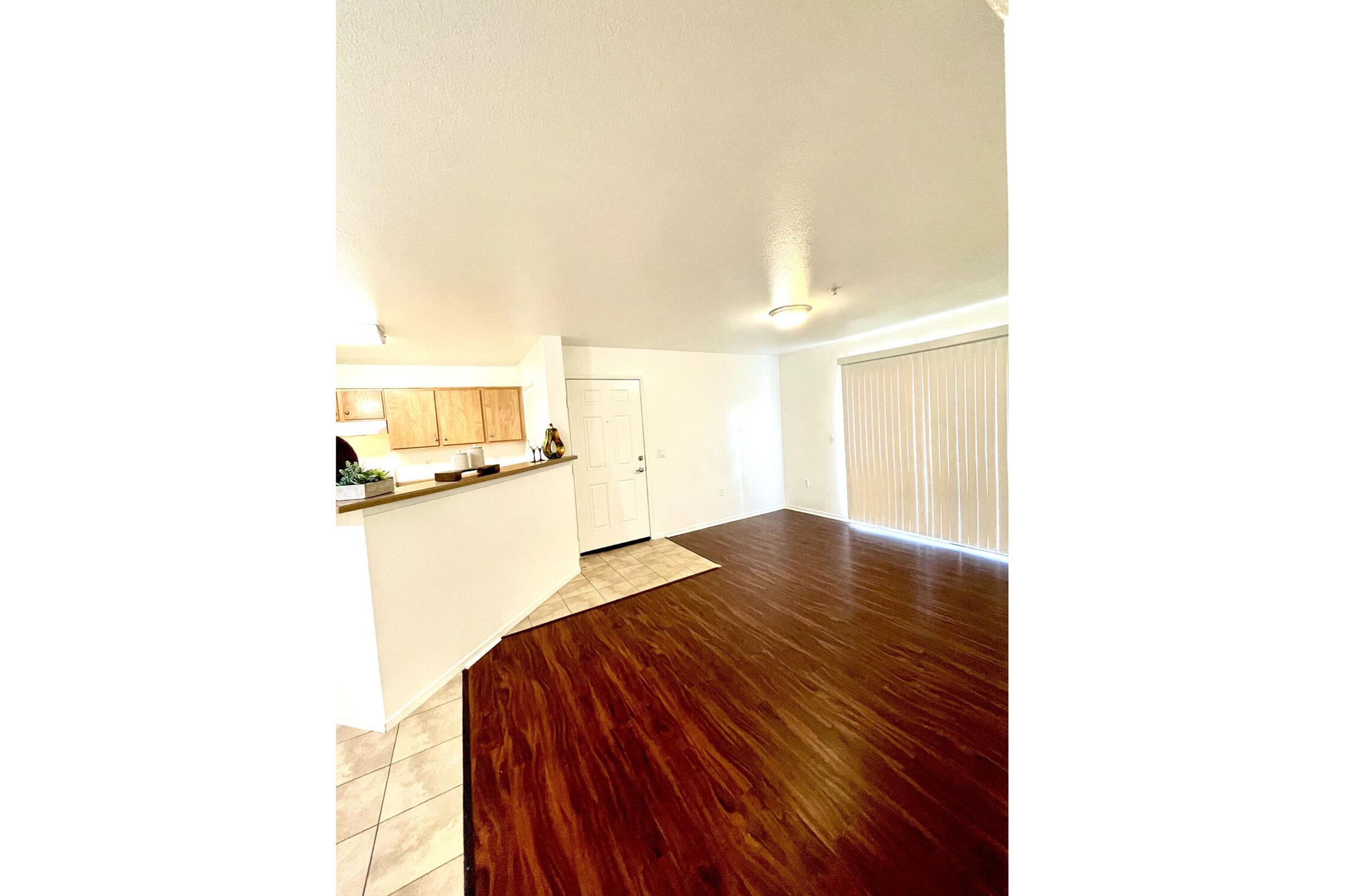 A spacious interior view of a living area with hardwood flooring and a light-colored wall. In the background, there is a kitchen with wooden cabinets and a countertop. A sliding glass door with blinds is visible, along with a small rug in the foreground. The space is well-lit and inviting.