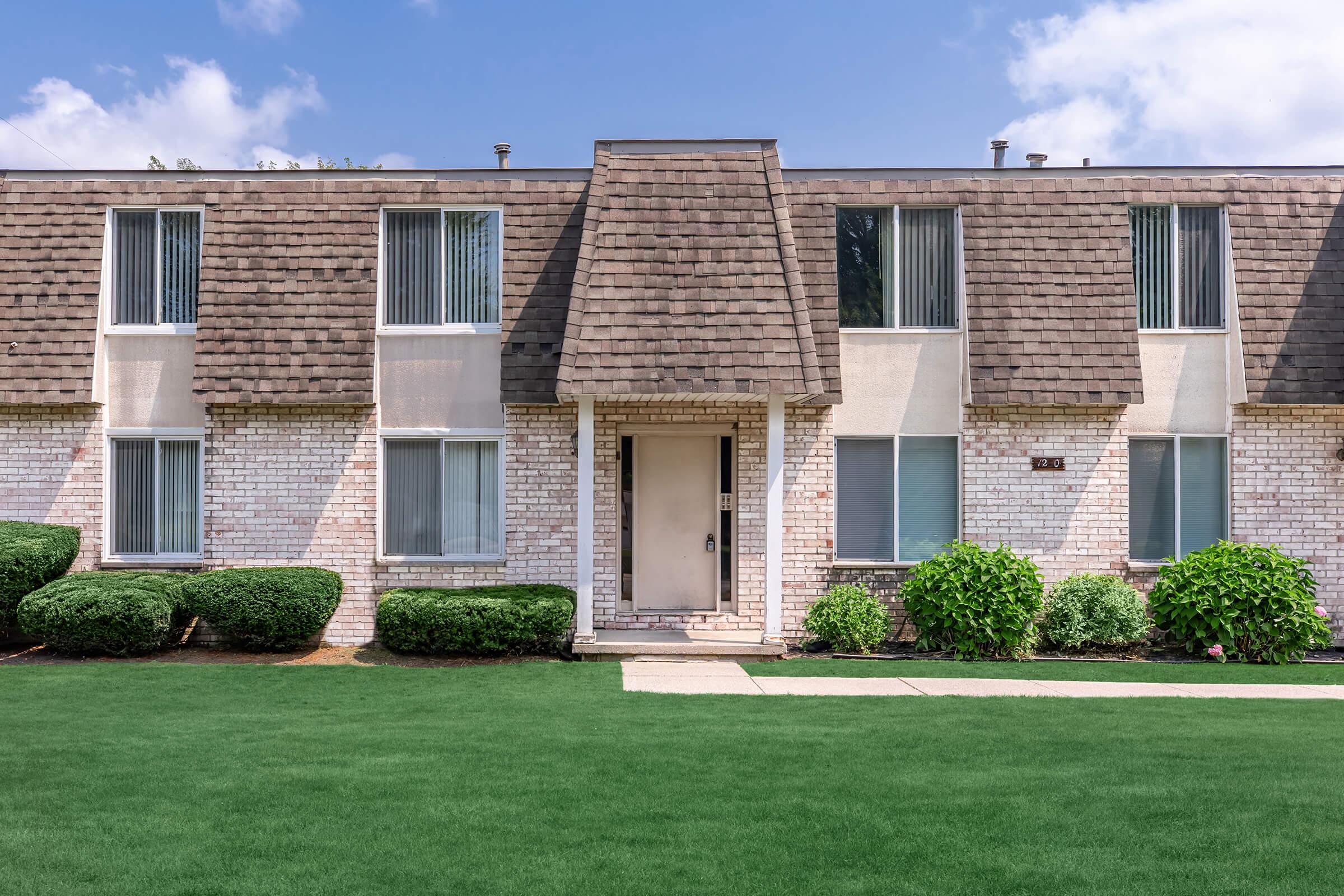 A two-story apartment building featuring a tan brick facade and a distinctive sloped roof. The front door is centrally located, flanked by large windows. Neatly trimmed green bushes line the walkway, which leads to a manicured lawn under a partly cloudy blue sky.