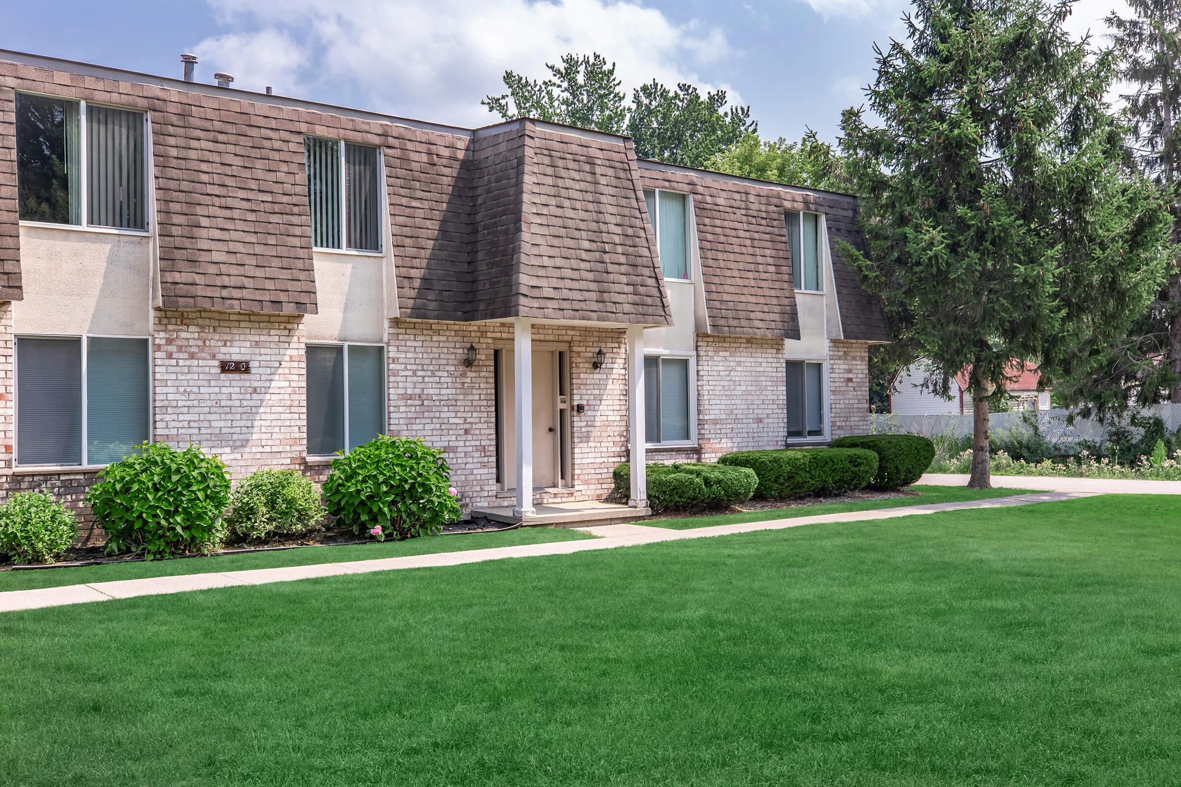 A well-maintained brick apartment building with a brown shingled roof, featuring multiple windows and a front door. The surrounding area includes neatly trimmed hedges and a lush green lawn under a partly cloudy sky.