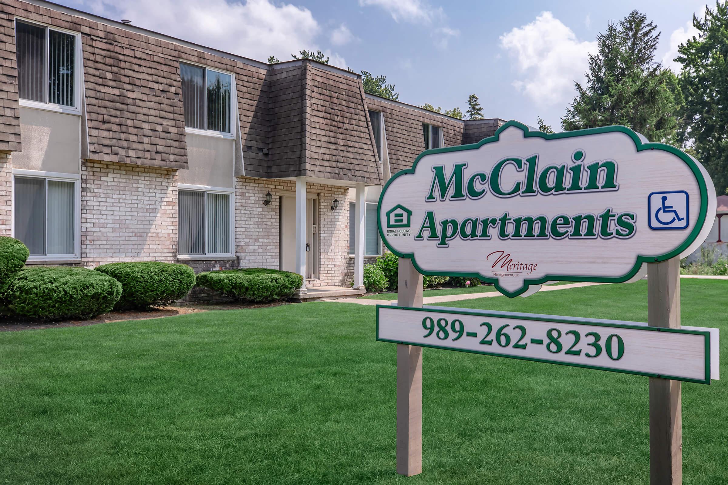 A sign for McClain Apartments featuring the phone number 989-262-8230, located in front of a two-story building with a sloped roof. The building has several windows and is surrounded by a well-maintained lawn and shrubs. The sign indicates accessibility features.