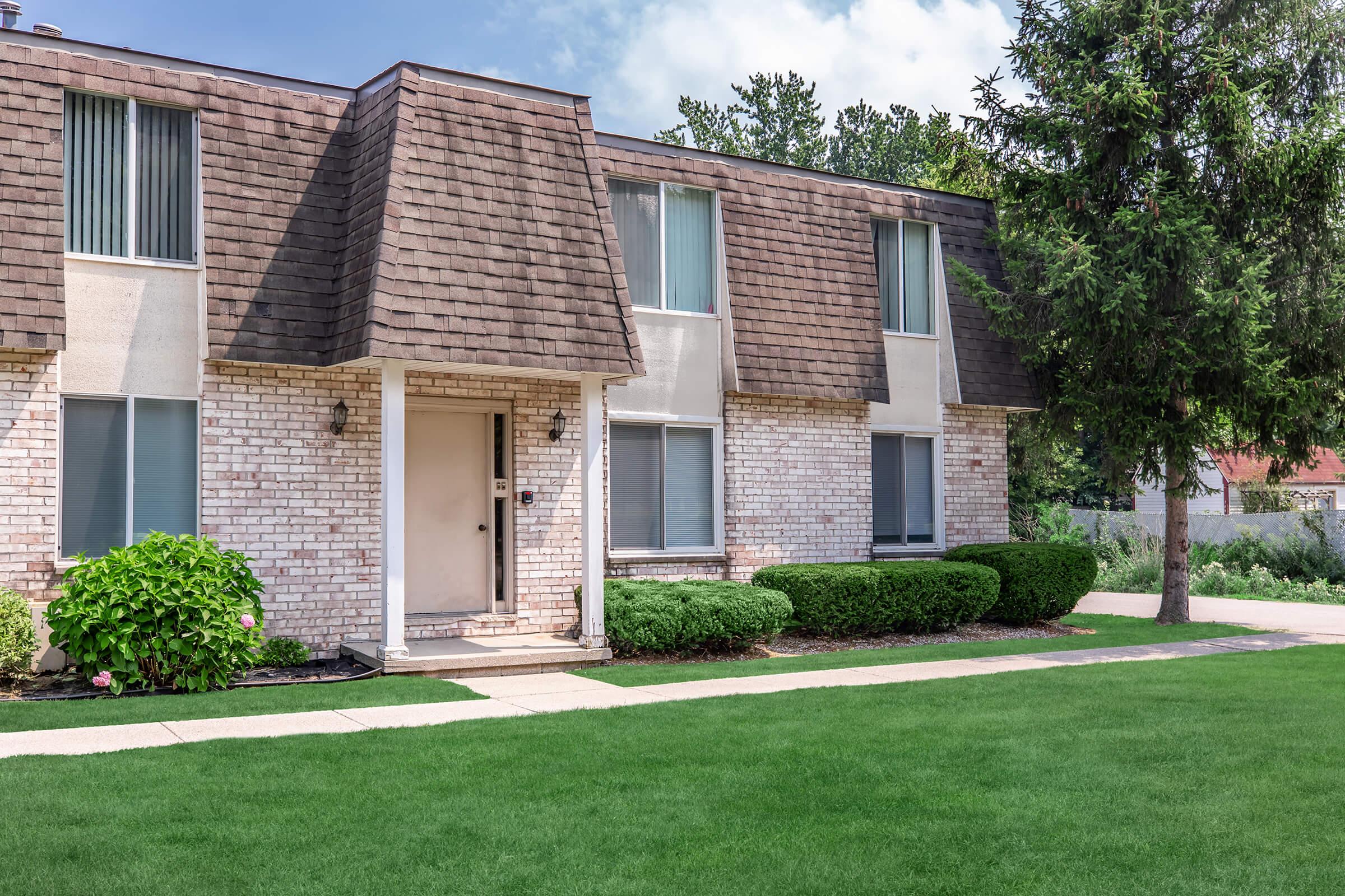A two-story brick apartment building with a sloped roof, featuring several windows. The entrance is framed by two shrubs on either side, and a well-manicured lawn is in front. Trees and shrubs are visible in the background under a clear blue sky.