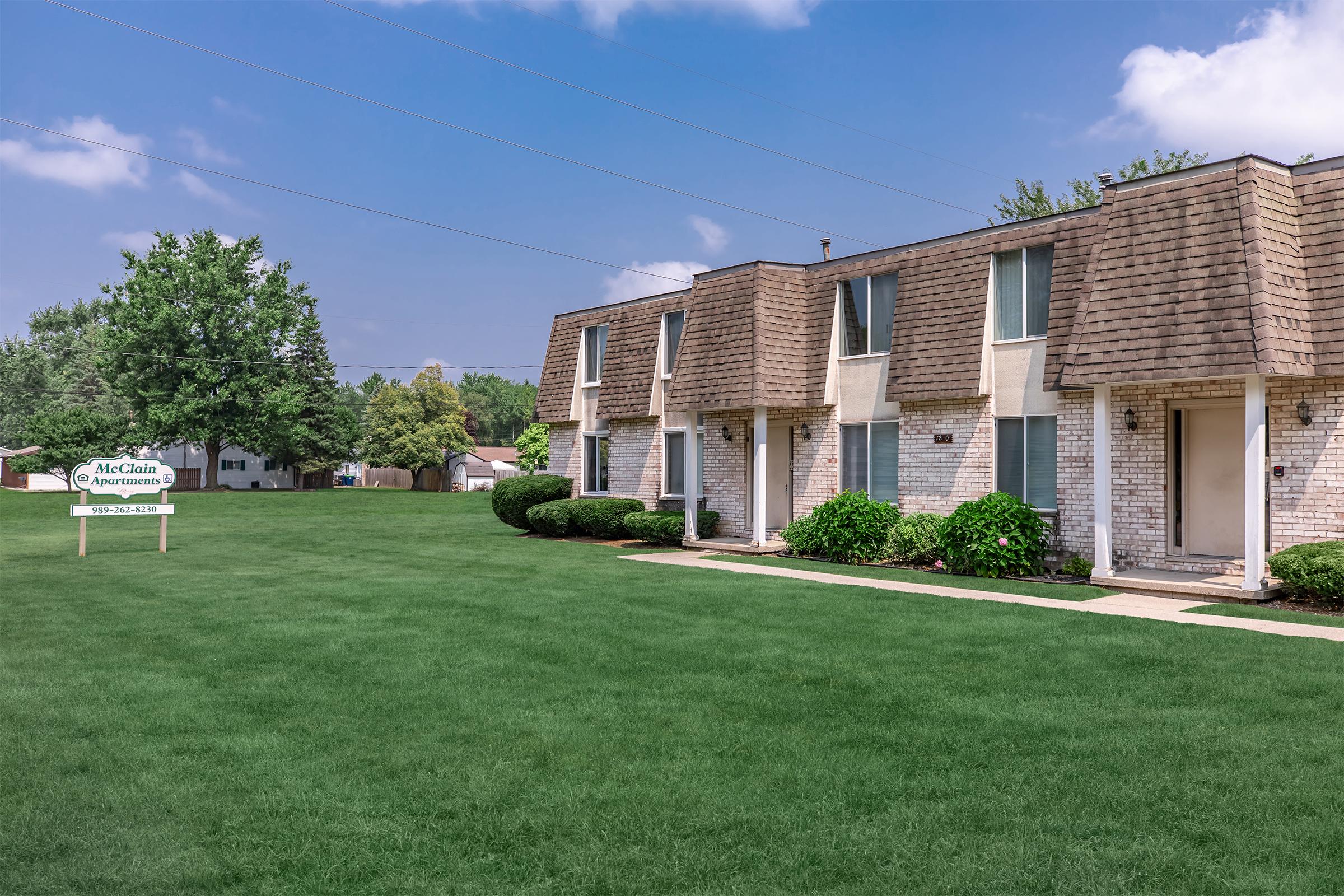 A view of a well-maintained apartment complex featuring two-story brick buildings with sloped roofs. The lush green lawn is neatly manicured, and there are small shrubs and bushes along the pathway. A sign for "McClain Apartments" is visible in the foreground. The sky is clear with a few clouds.
