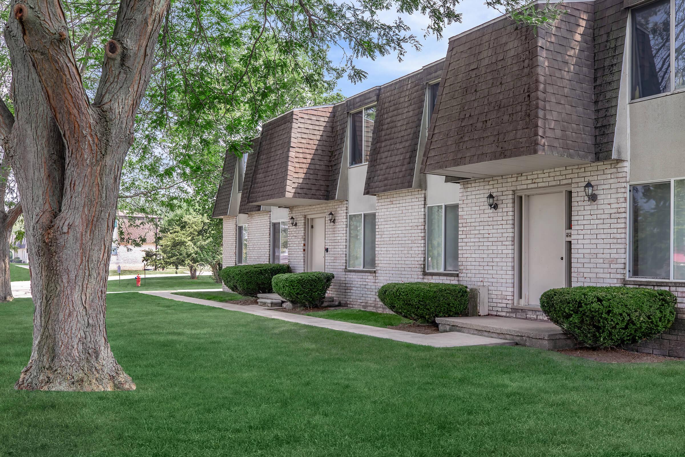 A row of modern townhouses with slanted roofs, featuring white brick exteriors and neatly trimmed shrubs. The grass is lush and green, and there are trees providing shade nearby. The scene is well-lit, suggesting a clear day. Sidewalks lead to individual entrances for each unit.