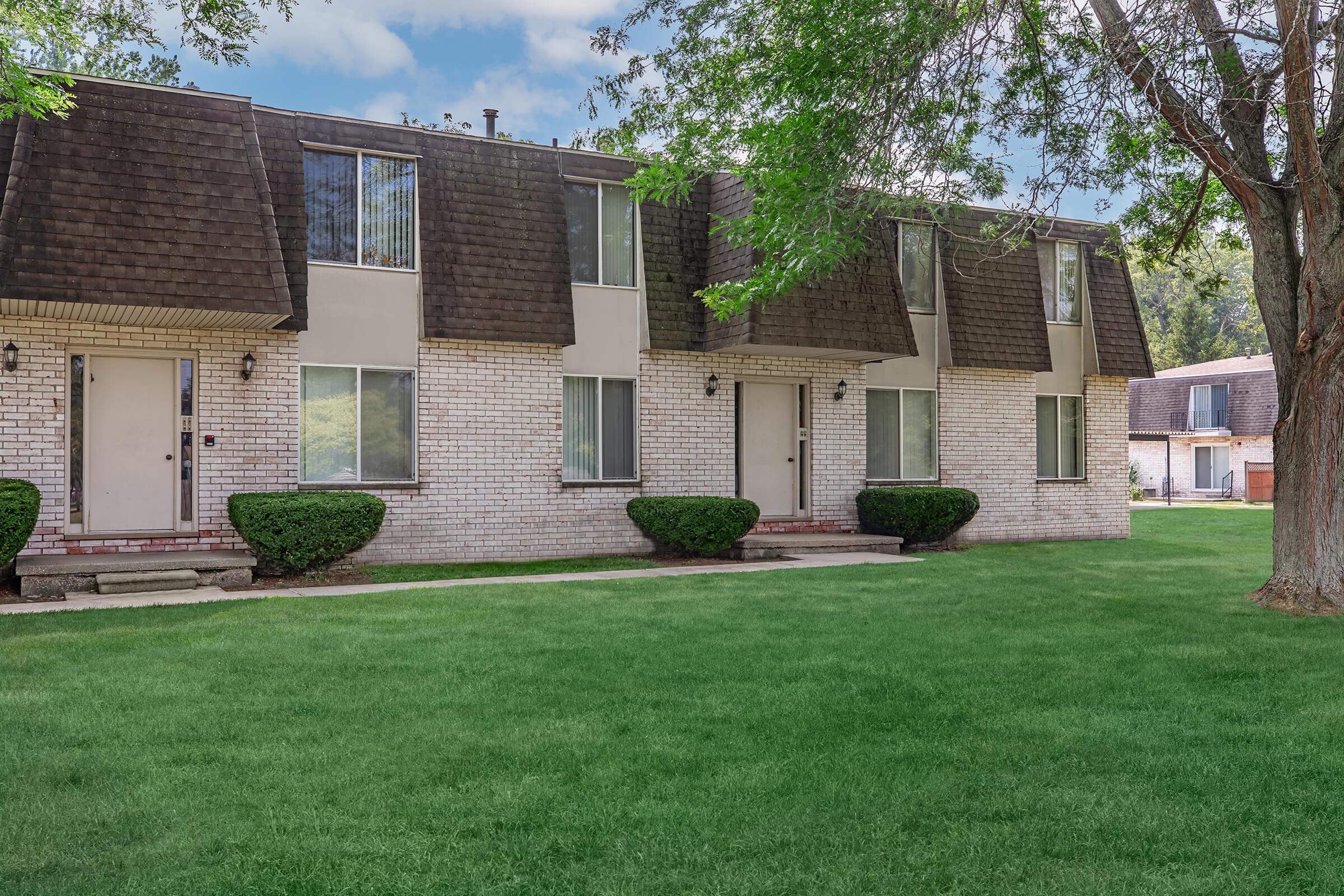 A brick apartment building with a brown shingled roof, featuring several windows and white metal doors. The building is set in a green lawn area with neatly trimmed shrubs on either side of the entrance. Trees provide shade around the property, with additional buildings visible in the background.