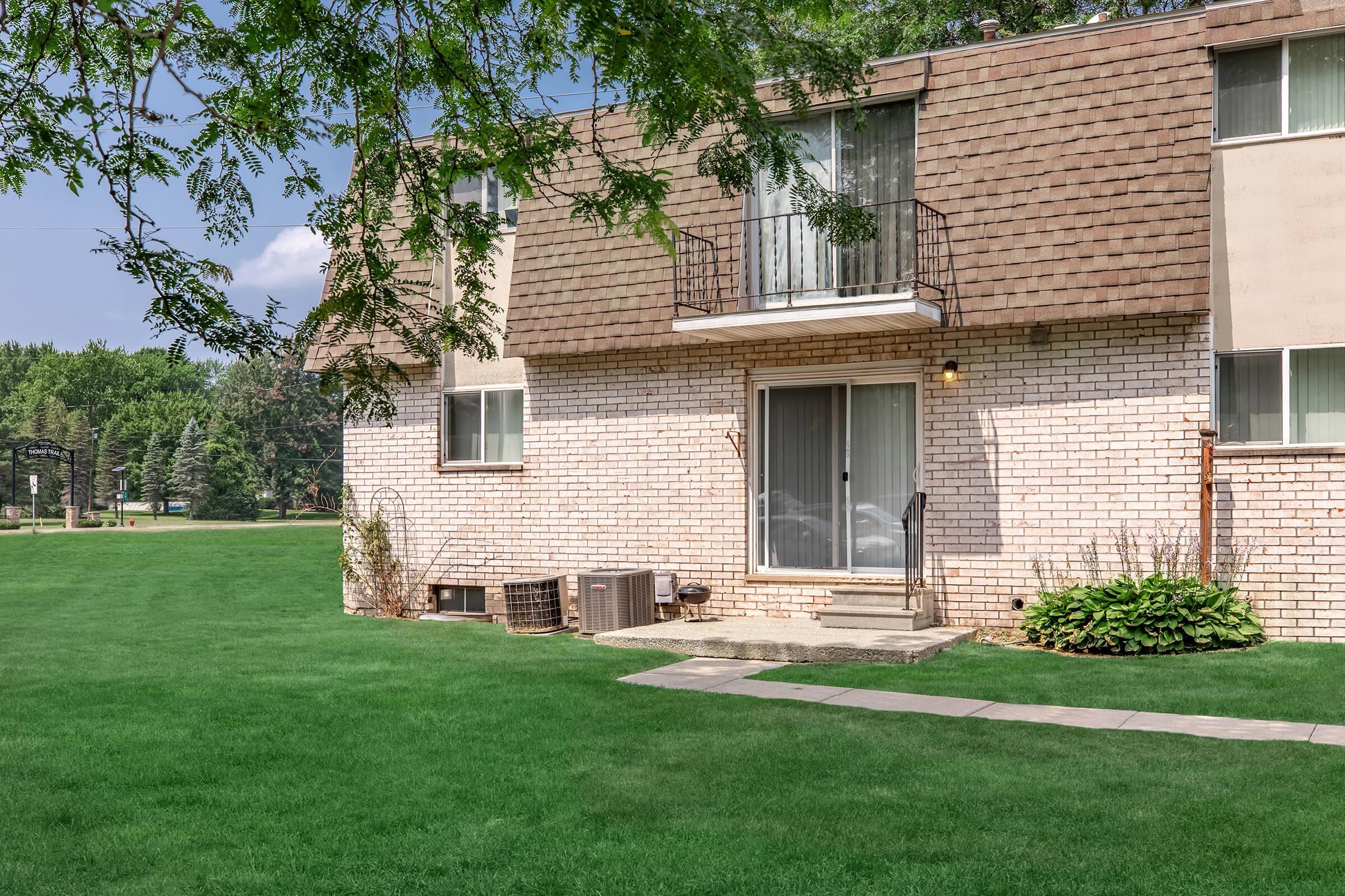 A two-story brick apartment building with a brown shingle roof. The ground level features a patio door with a small balcony above, and air conditioning units are visible on the side. The surrounding area is well-maintained grass, with trees in the background and a clear blue sky overhead.