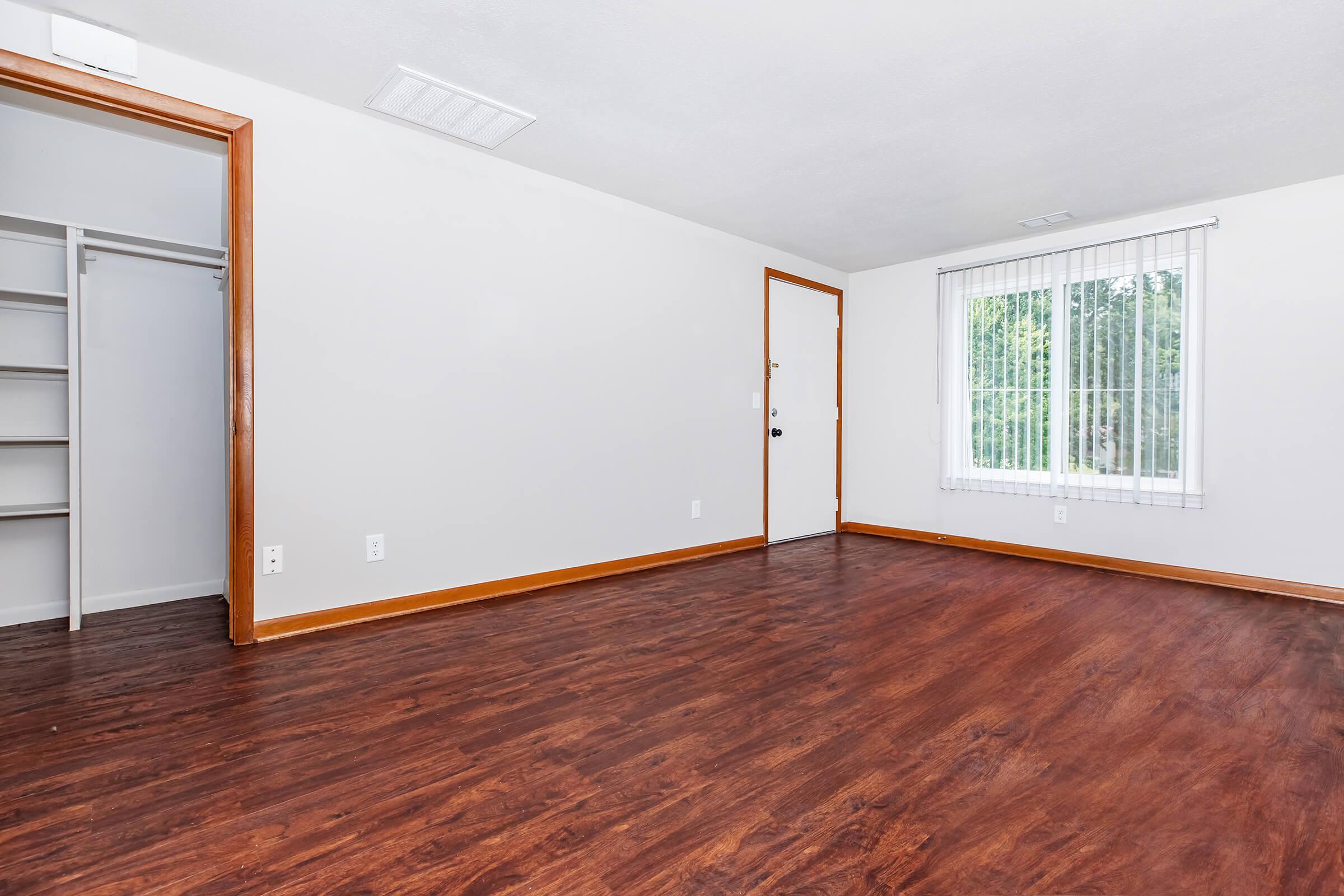 A spacious, empty room featuring hardwood-style floors, a bright window with white blinds, and a door leading outside. There is a closet with open shelves on one side, and the walls are painted a light neutral color. The overall ambiance is clean and inviting, suitable for various uses.