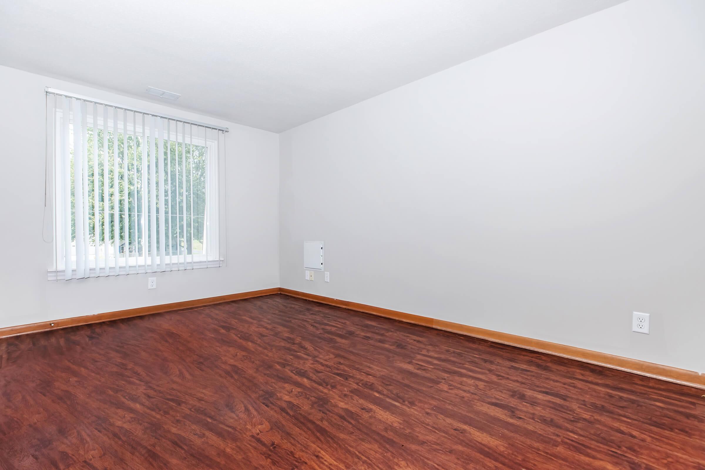 An empty room with light brown wooden flooring, a large window covered by white vertical blinds, and plain light gray walls. The room has a minimalistic appearance and no furniture. A small white wall outlet is visible on the right side.
