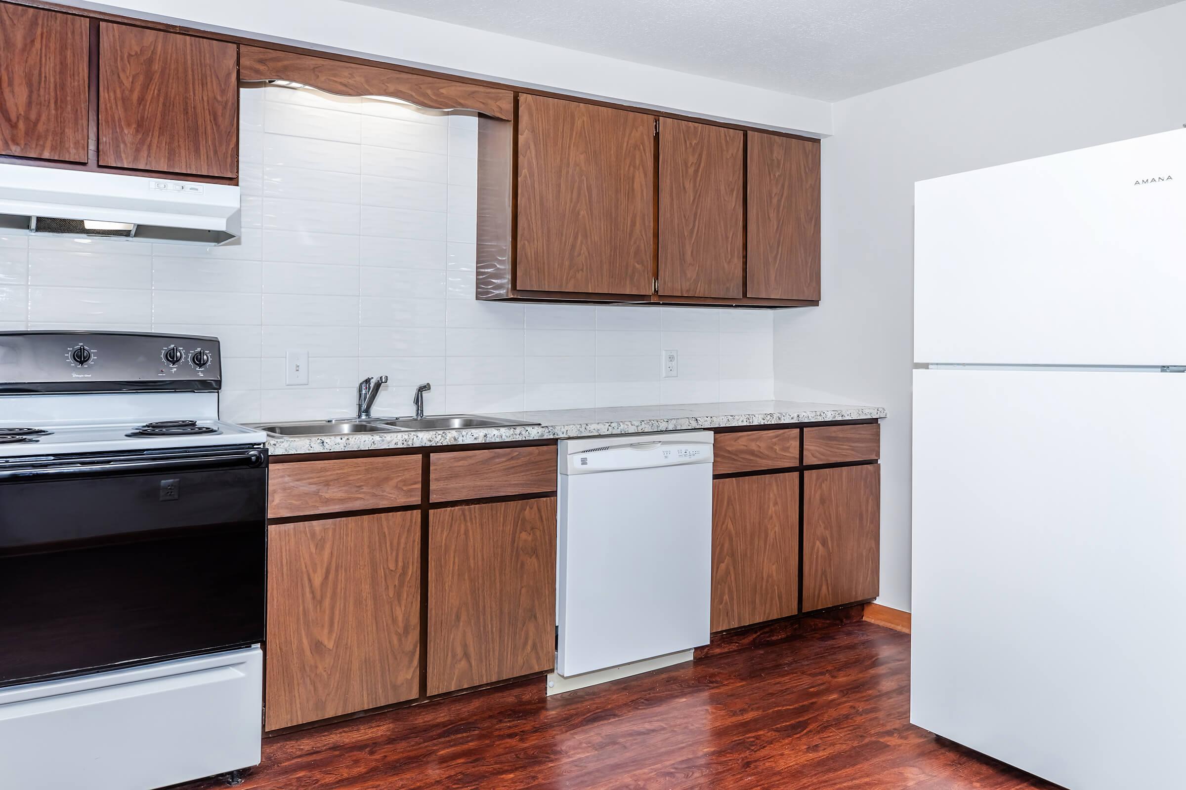 Modern kitchen featuring wooden cabinets, a sink, an oven, and a dishwasher. The countertops are made of granite, and there is a white refrigerator to the right. The flooring is dark wood, and the wall is painted in a light color, providing a bright and clean appearance.