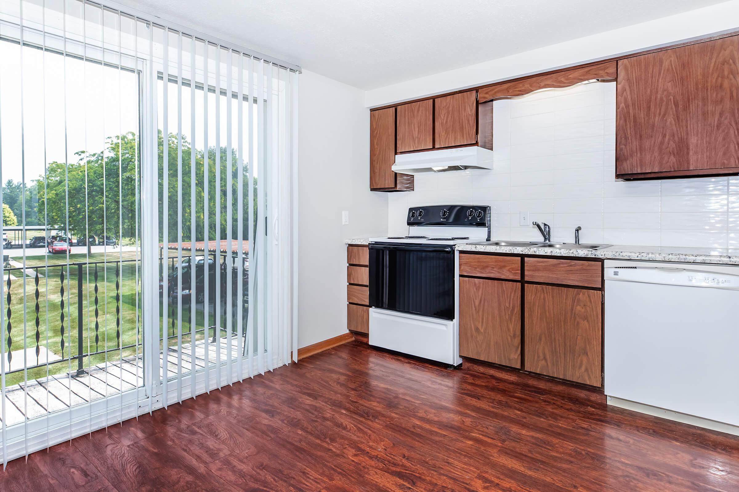 A modern kitchen featuring brown wooden cabinets, a black stove, and a white refrigerator. The area has a sliding glass door that opens to a balcony, letting in natural light, and has a wood-like floor for a cozy ambiance.