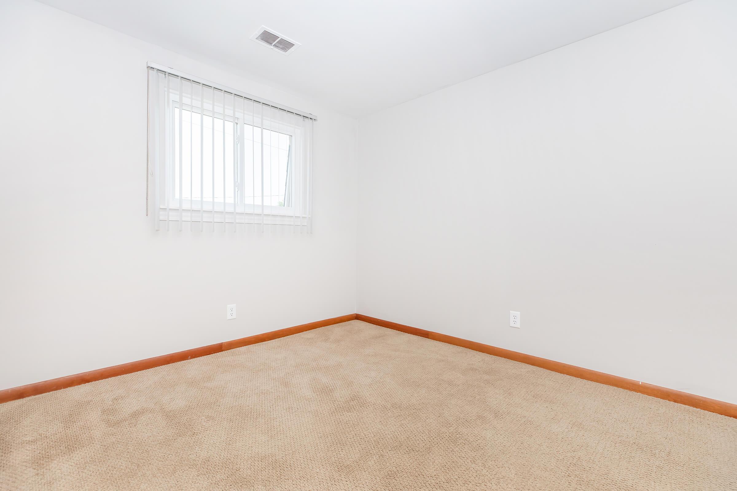 A vacant room with beige carpet and white walls. A window with blinds is on one wall, allowing natural light to enter. There are no furniture or decorations present, creating a minimalist and empty appearance.