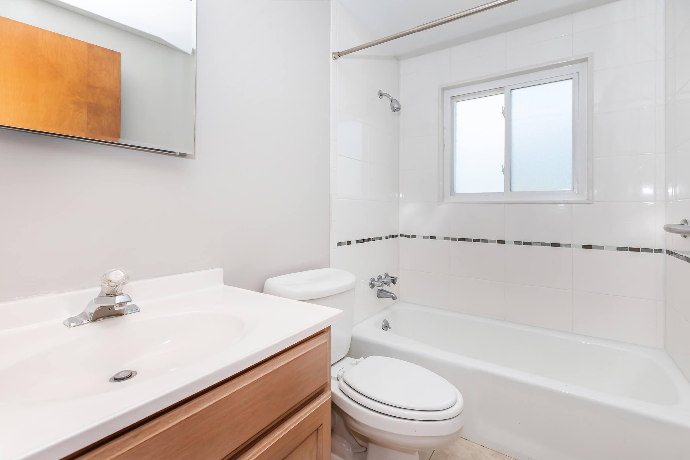 A clean, modern bathroom featuring a white bathtub, a sink with a wooden cabinet, and a toilet. There is a window providing natural light, and the walls are tiled in white with a decorative border. A mirror is mounted above the sink.