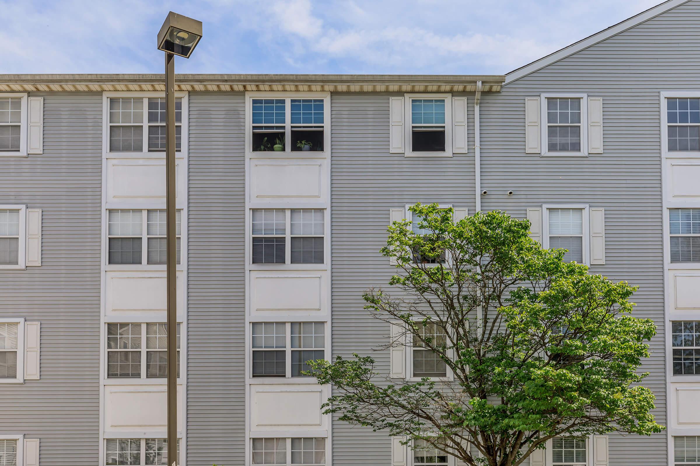 A multi-story residential building with gray siding features several windows, some of which are open. A lamp post stands beside the building, and a small tree is visible in the foreground, providing greenery against the backdrop of the structure and blue sky.