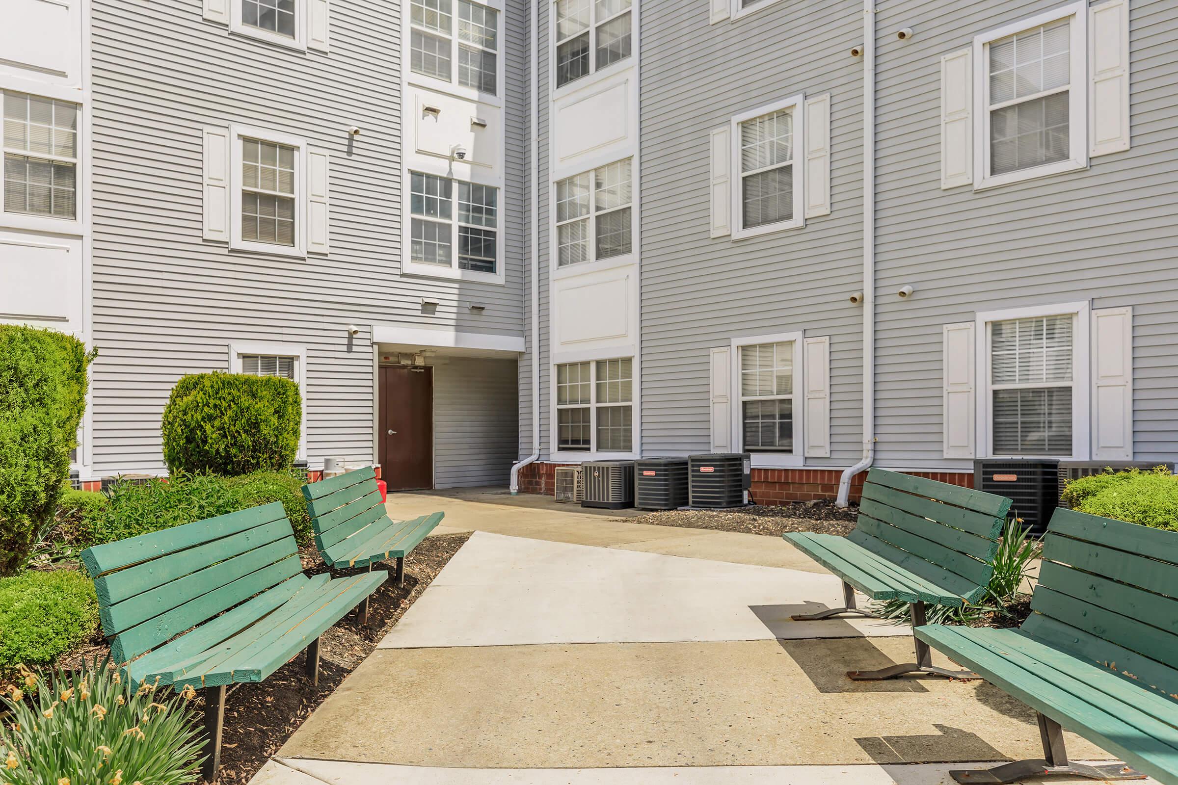 A courtyard area featuring two green benches facing a concrete walkway. Surrounding the benches are small shrubs and plants, with a backdrop of a multi-story gray apartment building that has white window shutters and air conditioning units visible. The environment appears well-maintained and inviting.