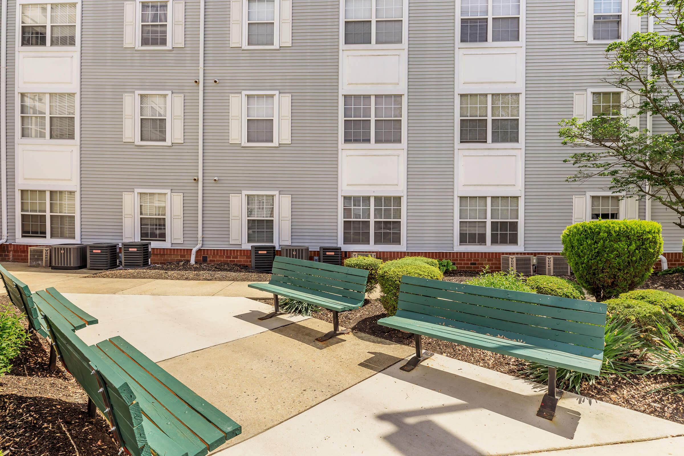 Two green benches positioned along a paved path in a landscaped area, with shrubs and decorative plants. In the background, a multi-story building with gray siding and white windows is visible. Air conditioning units are placed near the building's foundation.