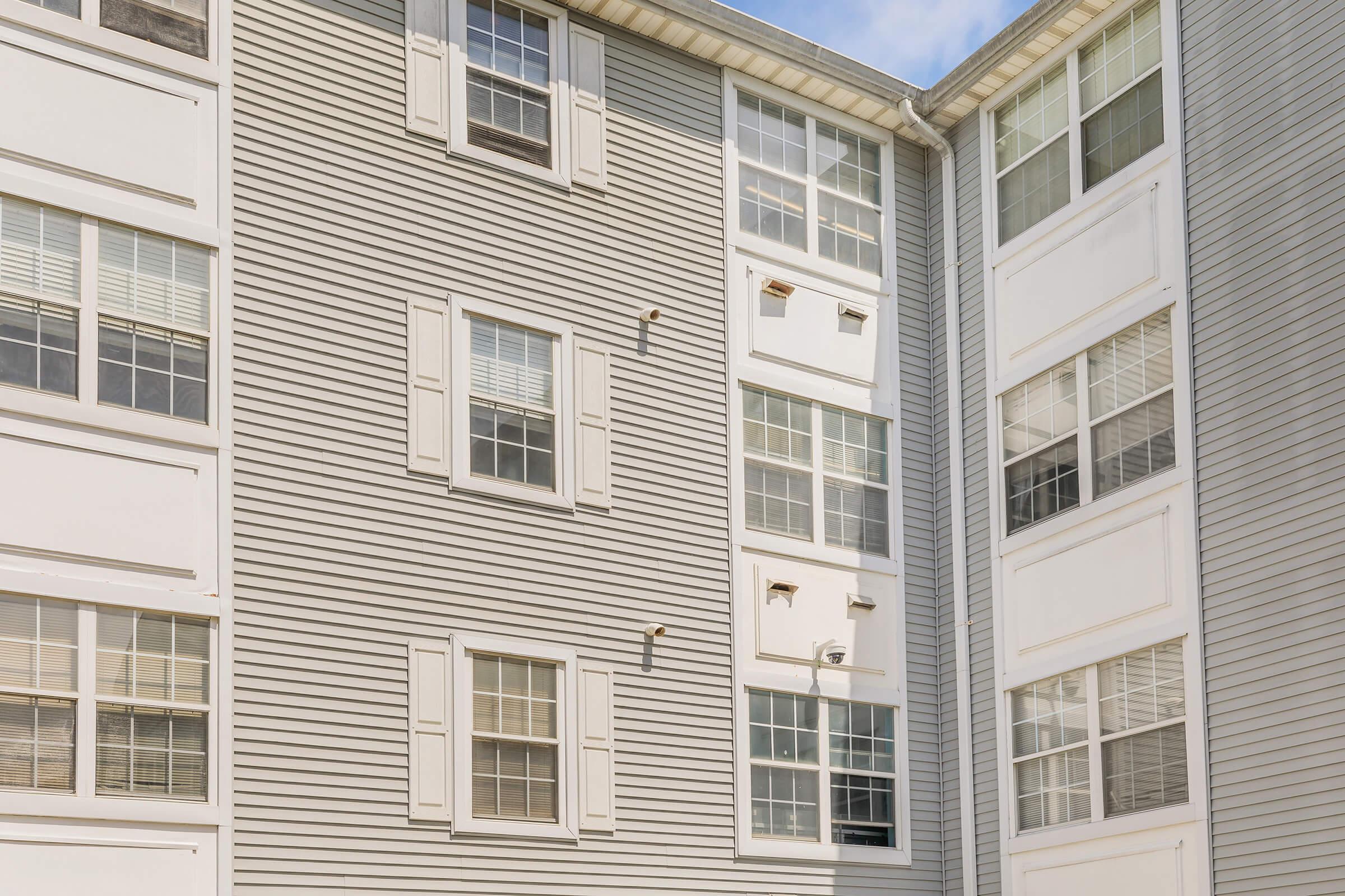 Exterior view of a multi-story apartment building featuring multiple windows with white shutters. The facade is clad in grey siding, showcasing a modern architectural design. Bright sunlight illuminates the building, highlighting its clean lines and contemporary look against a blue sky.