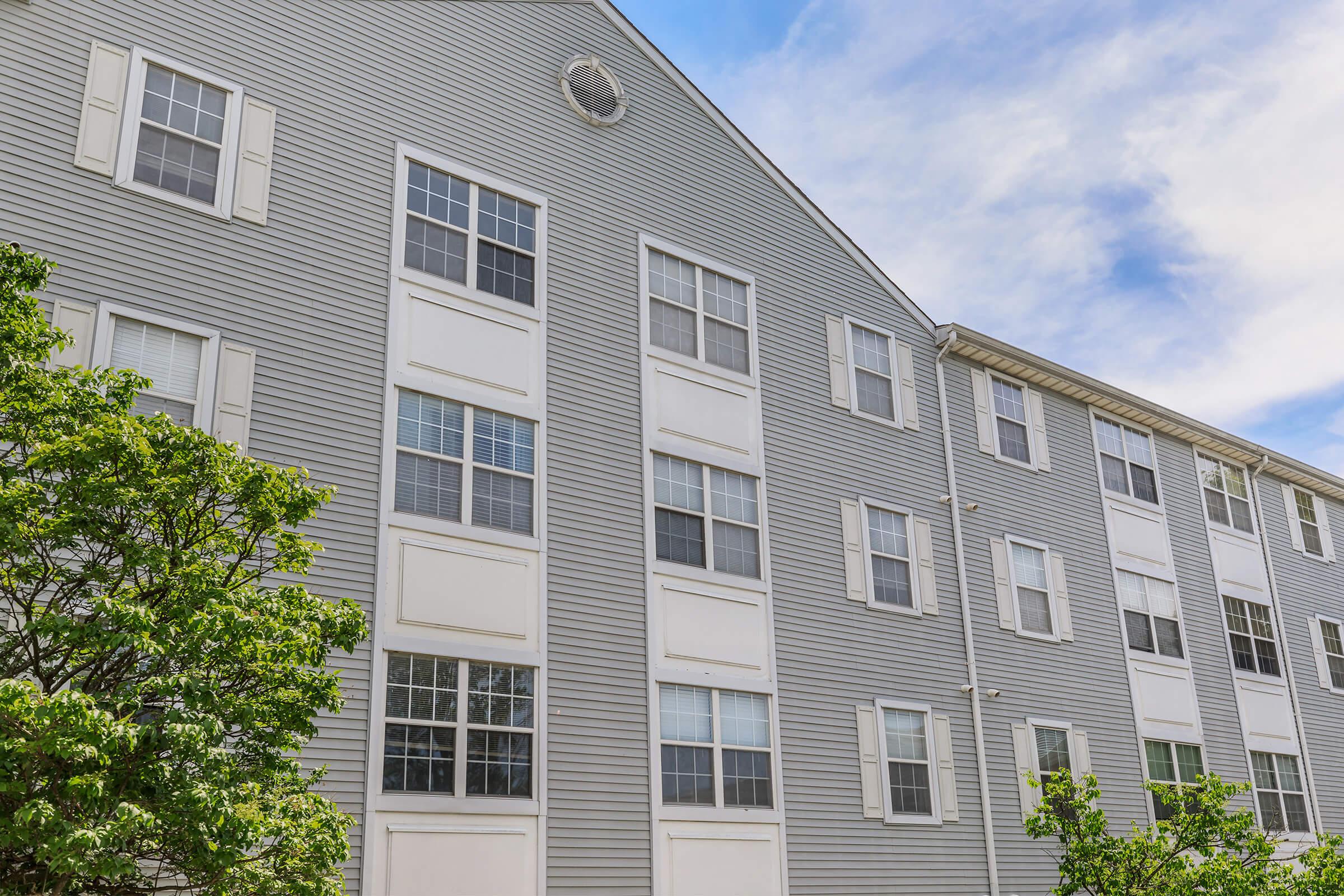 Gray multi-story apartment building with multiple windows and white shutters, surrounded by greenery. The sky is partly cloudy, adding a bright atmosphere to the scene.