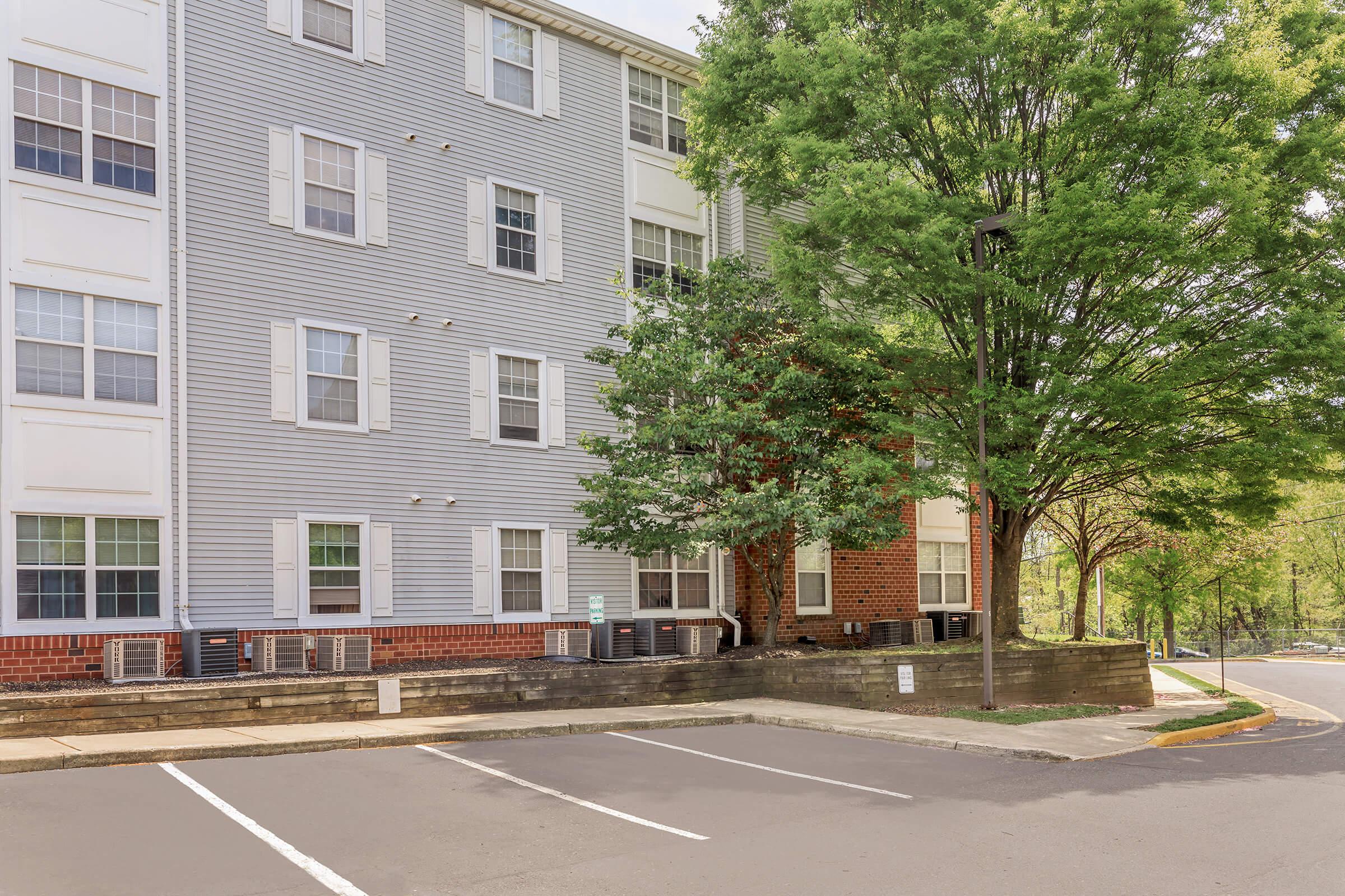 A three-story apartment building with a combination of gray and red siding, featuring multiple windows and air conditioning units. Green trees and shrubs are visible in front, with a small parking area in the foreground. The setting appears to be suburban and well-maintained.