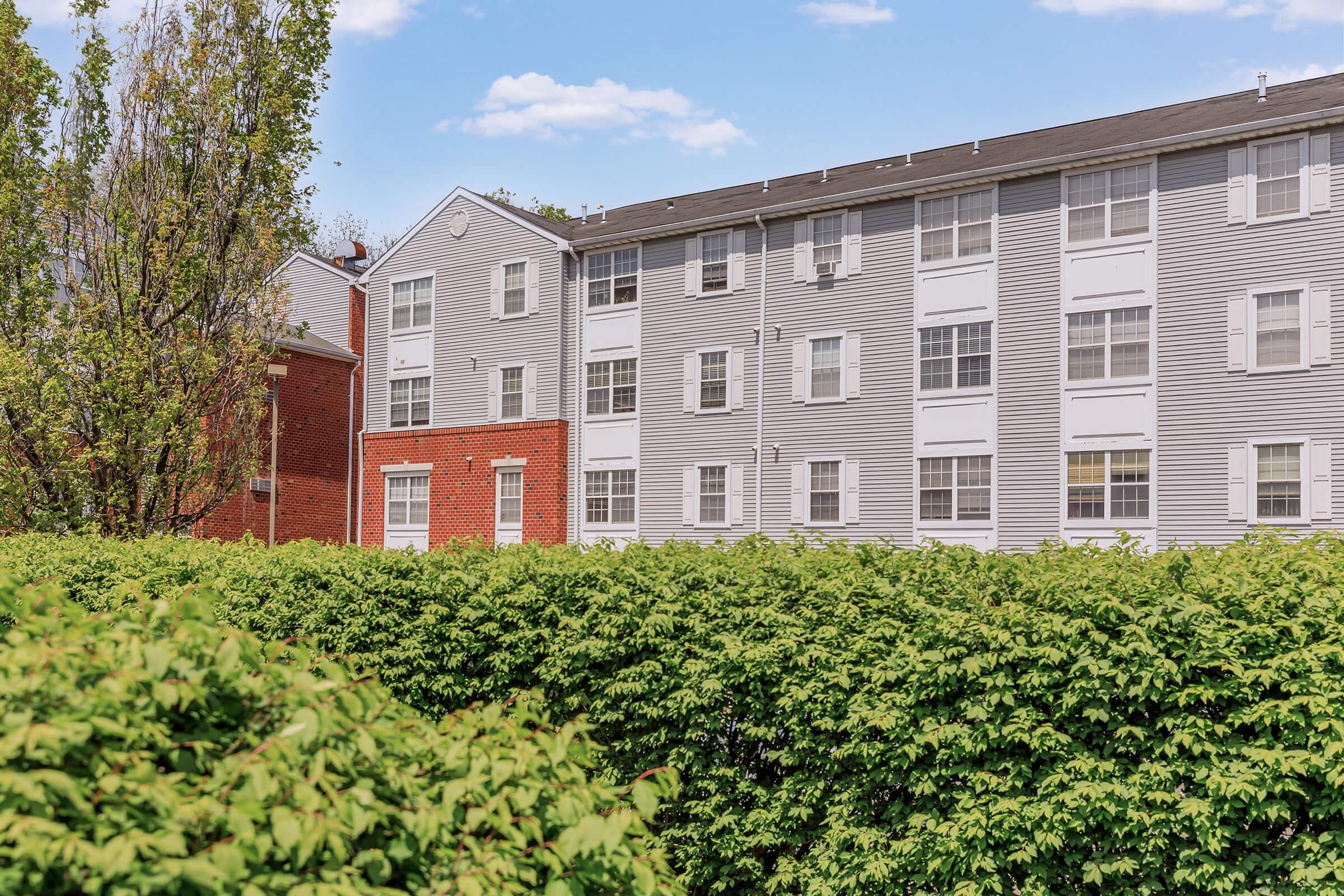 View of a residential apartment complex featuring a mix of gray and red brick buildings. The lower part of the image shows lush green hedges, creating a natural border. In the background, there are blue skies with scattered clouds, enhancing the outdoor setting.