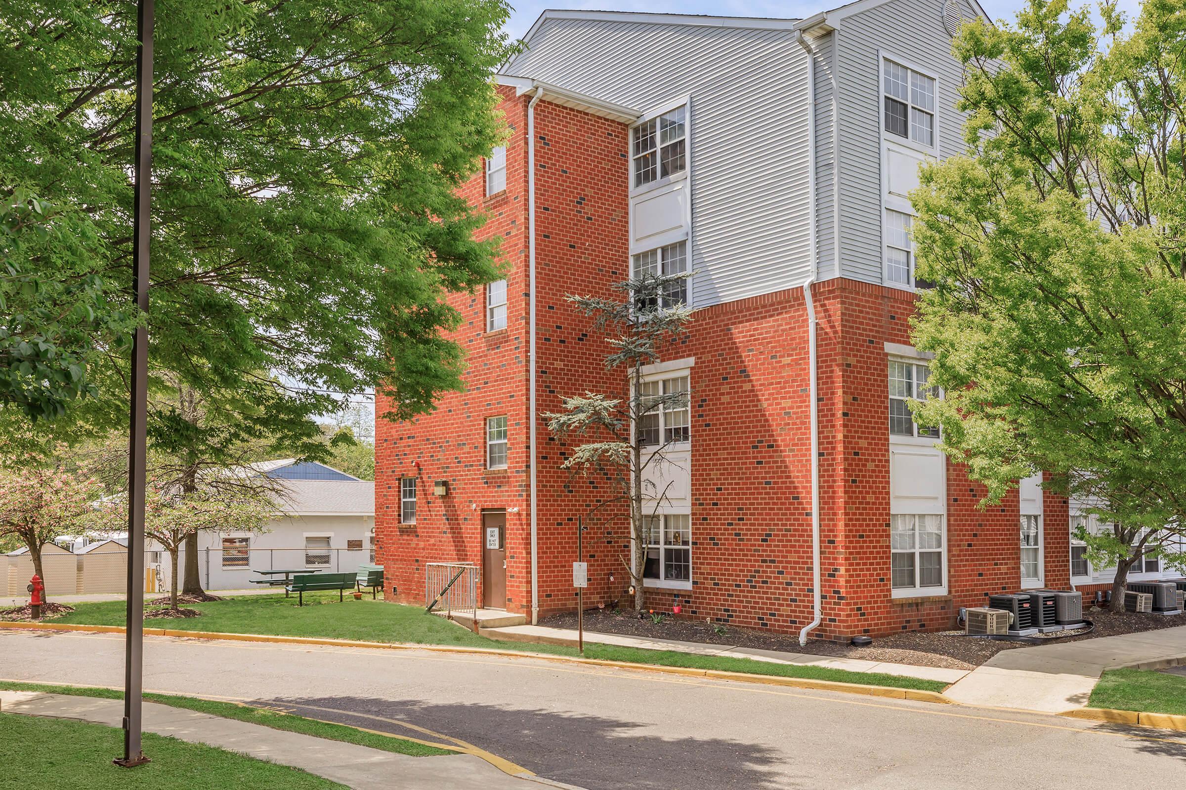 A modern, three-story brick building with light gray siding and large windows, surrounded by green trees. A paved road curves around the building, leading to a nearby structure in the background. The scene is bright and well-maintained, highlighting a peaceful residential area.