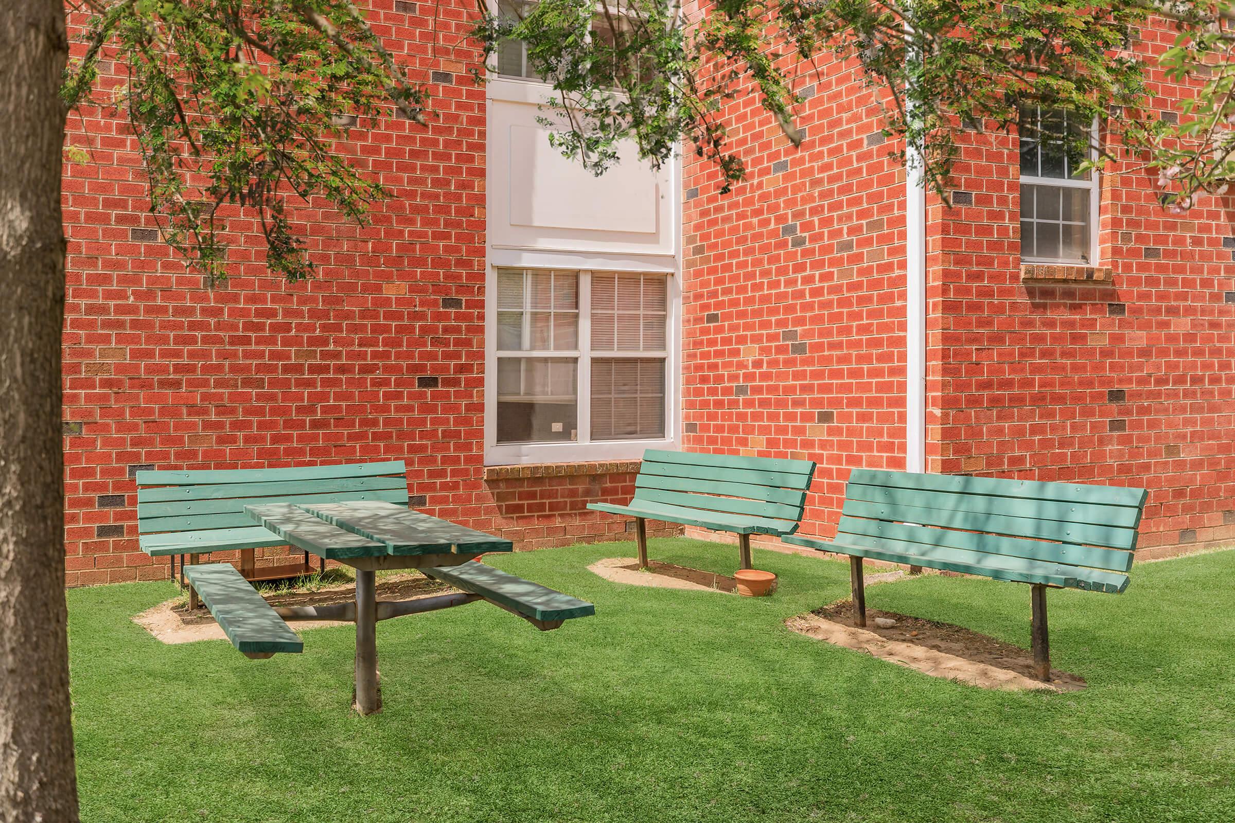 Outdoor seating area featuring green benches and a table on a grassy patch, set against a backdrop of a red brick wall. Sunlight illuminates the scene, with a tree providing some shade. Ideal for relaxation or casual meetings.