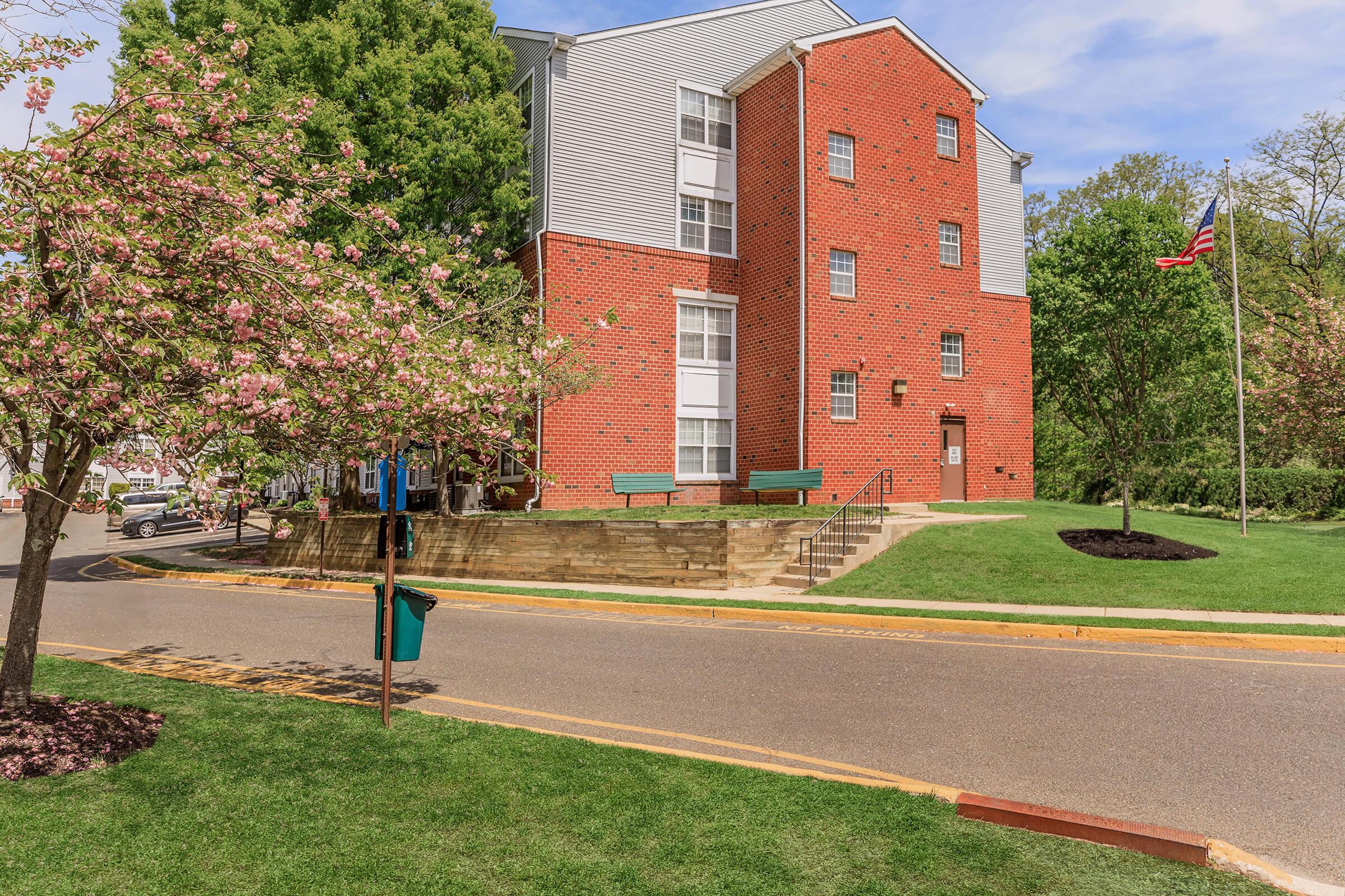 A brick building with multiple stories and large windows, surrounded by green grass and trees. Pink flowering trees are in bloom nearby, and a flagpole displaying the American flag is visible. A sidewalk leads to the building entrance, which has stone steps and a bench in front.