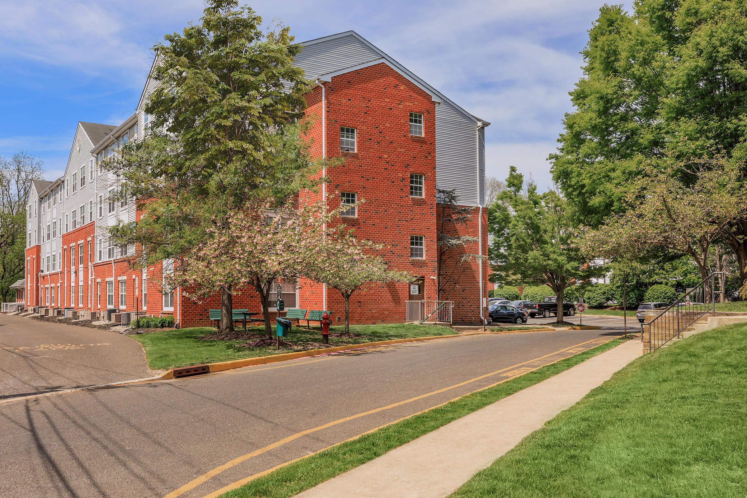 A red brick apartment building with multiple floors, surrounded by green trees and landscaped areas. A paved road runs alongside the building, and there's a sidewalk with a bench visible. The scene is bright with clear blue skies and a few parked cars in the background.