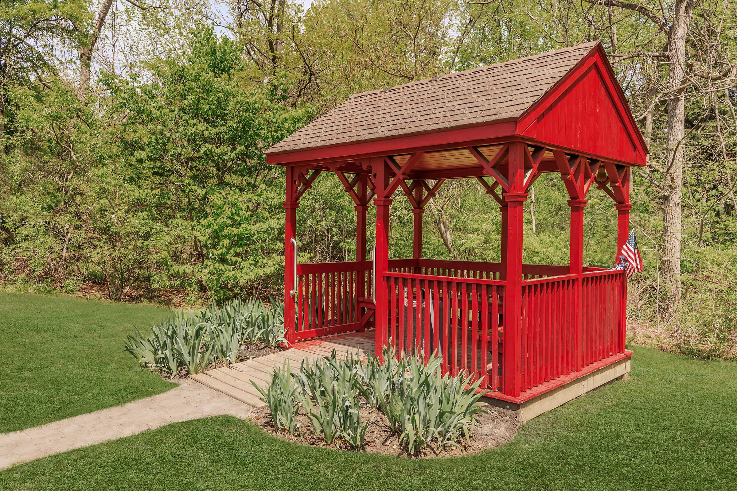 A bright red gazebo surrounded by lush greenery. The gazebo features a peaked roof, open sides, and is situated on a pathway leading through well-maintained grass and garden beds with blooming flowers. A small American flag decorates one corner, adding charm to the peaceful outdoor setting.