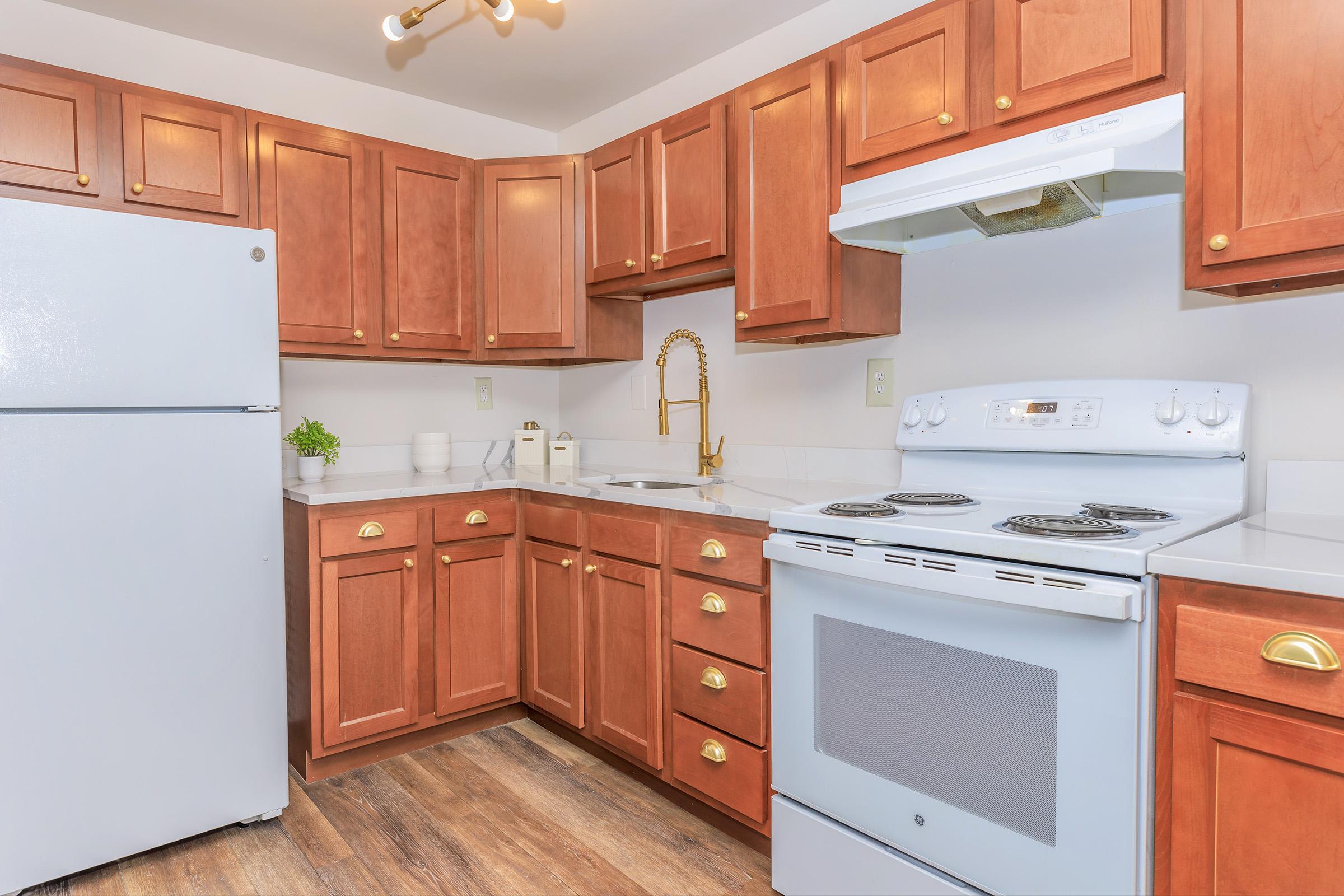 A modern kitchen featuring wooden cabinets, a white stove with an oven, and a matching refrigerator. There is a stainless steel sink with a gold faucet, and the countertop is white with minimal decorative items. The floor is made of wood, providing a warm and inviting atmosphere.