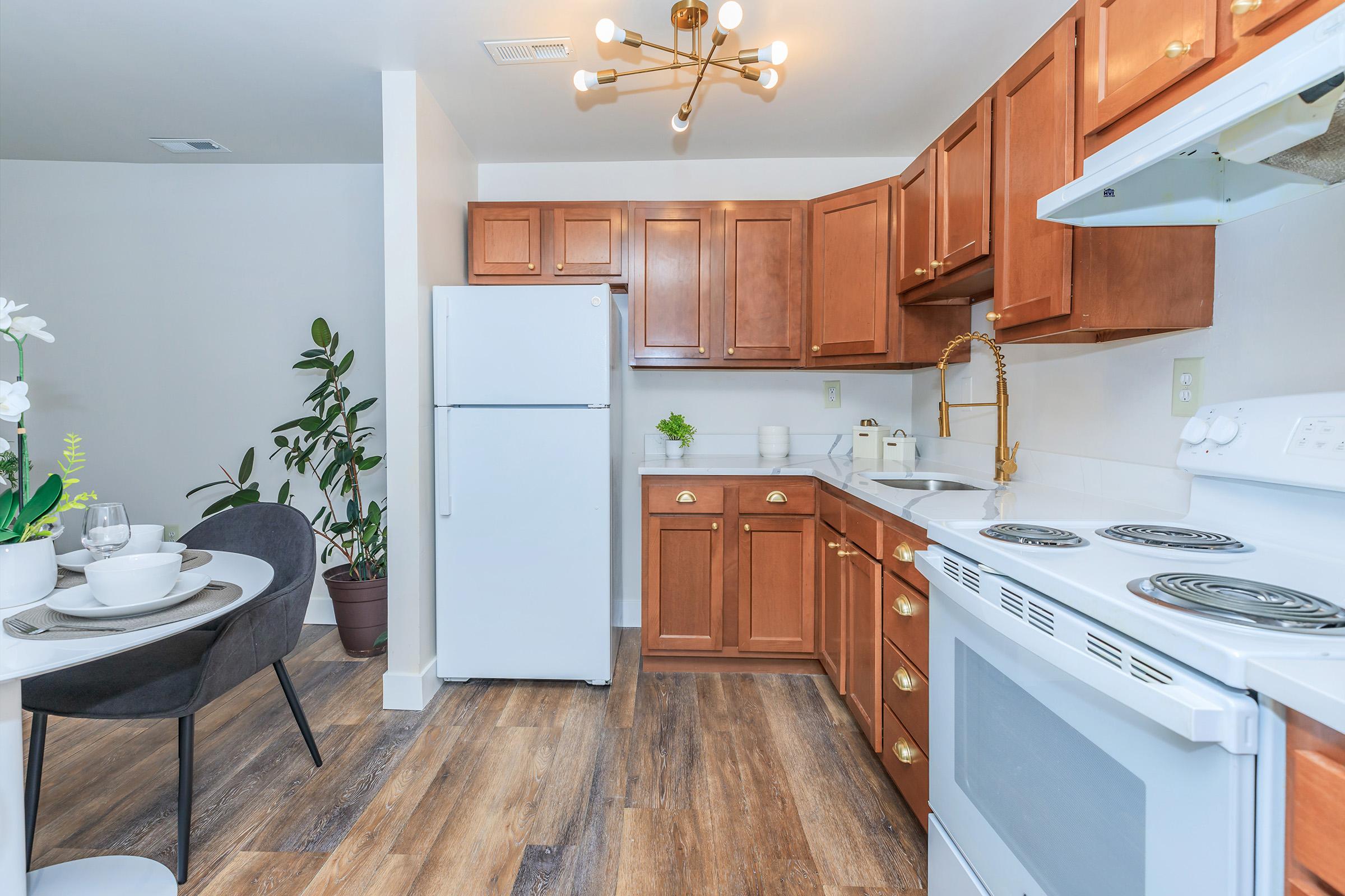 A modern kitchen featuring wooden cabinets, a white stove, and refrigerator. There is a dining area with a round table set for two, and indoor plants add a touch of greenery. The flooring is wood-like, and the overall color scheme is bright and inviting.