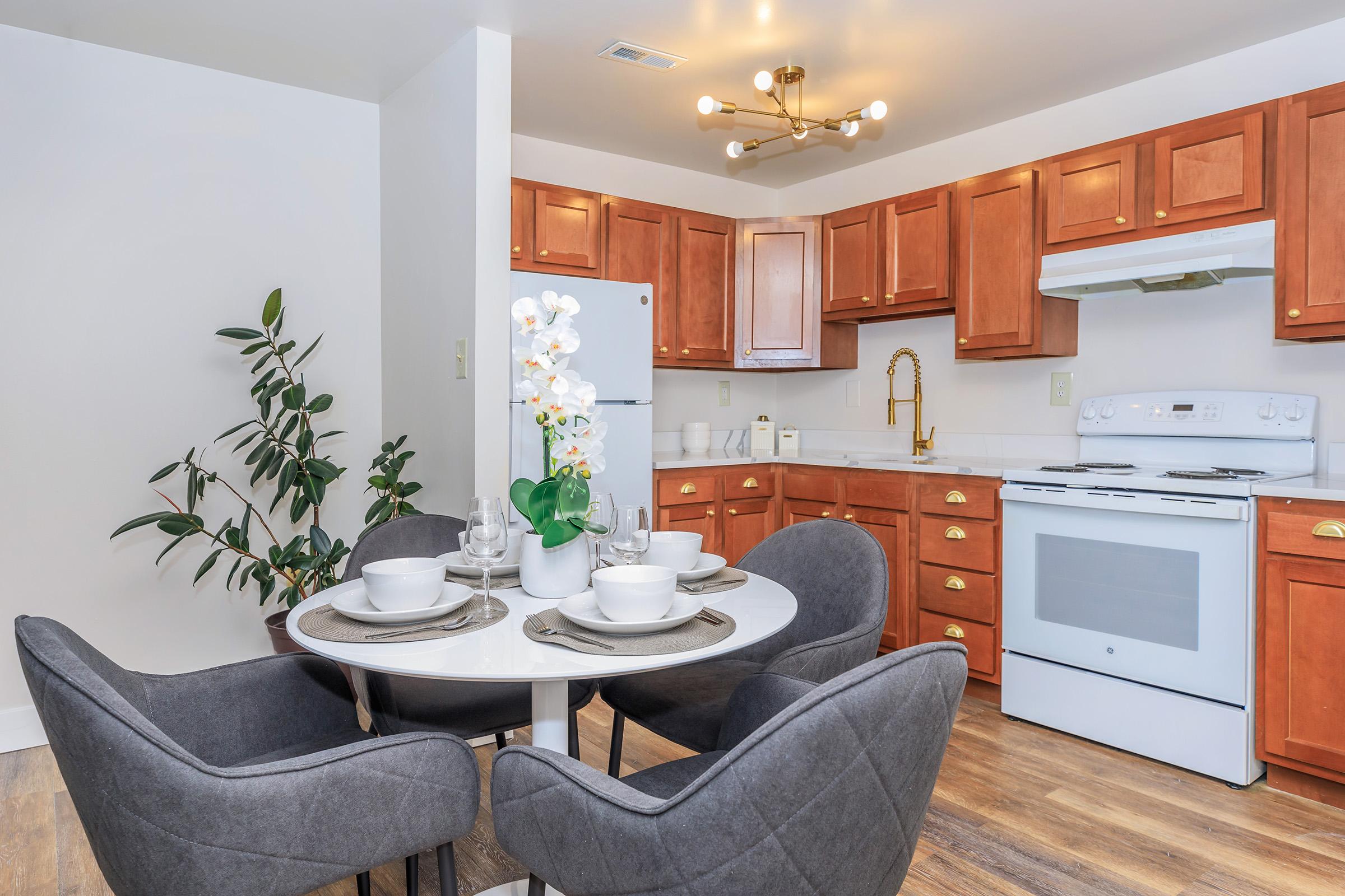 Modern kitchen featuring wooden cabinets, a white stove, and a round dining table with gray chairs. A decorative plant and a vase with white orchids adorn the table, creating a welcoming atmosphere. Bright lighting enhances the space, giving it a warm and inviting feel.