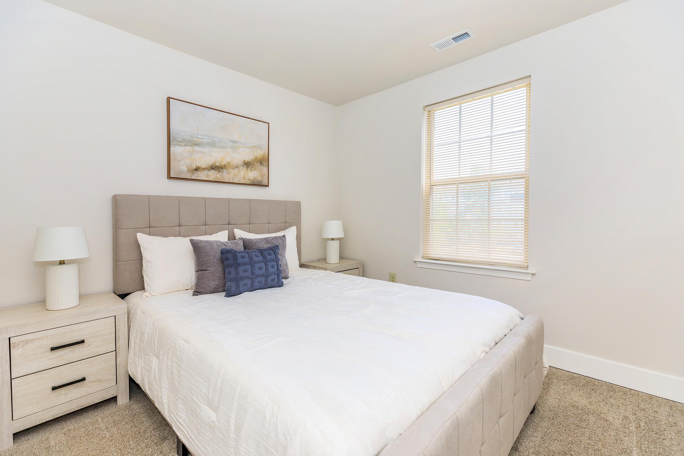 A cozy bedroom featuring a light-colored bed with white linens and a decorative blue pillow. There are two bedside tables with lamps, and a window with blinds allows natural light to fill the space. A simple landscape painting adorns the wall above the bed, adding a touch of decor.