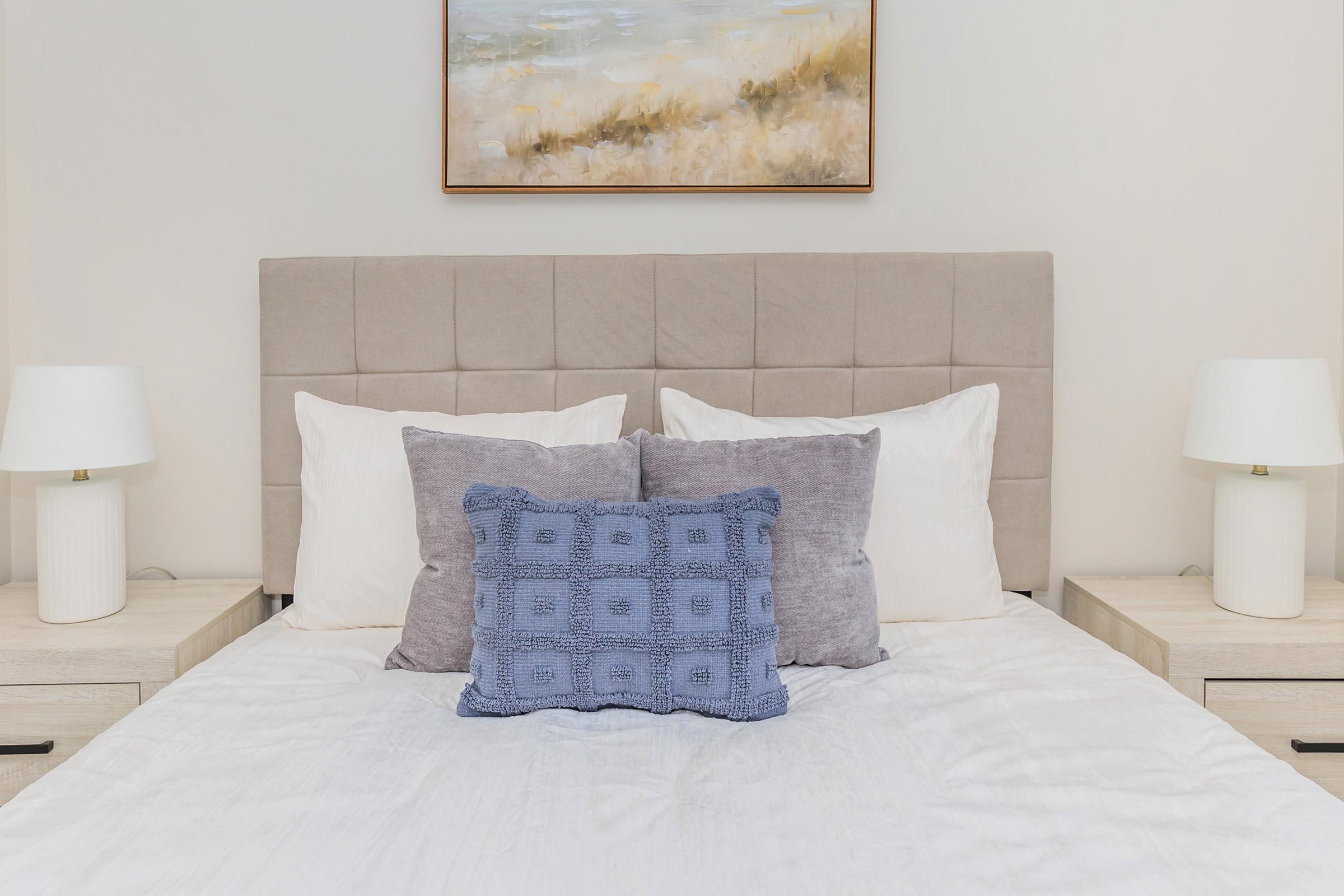 A neatly made bed with a light-colored comforter, featuring several decorative pillows in shades of white and blue. On either side of the bed, there are stylish nightstands with modern lamps. Above the bed, there is a framed abstract painting that complements the soft color palette.