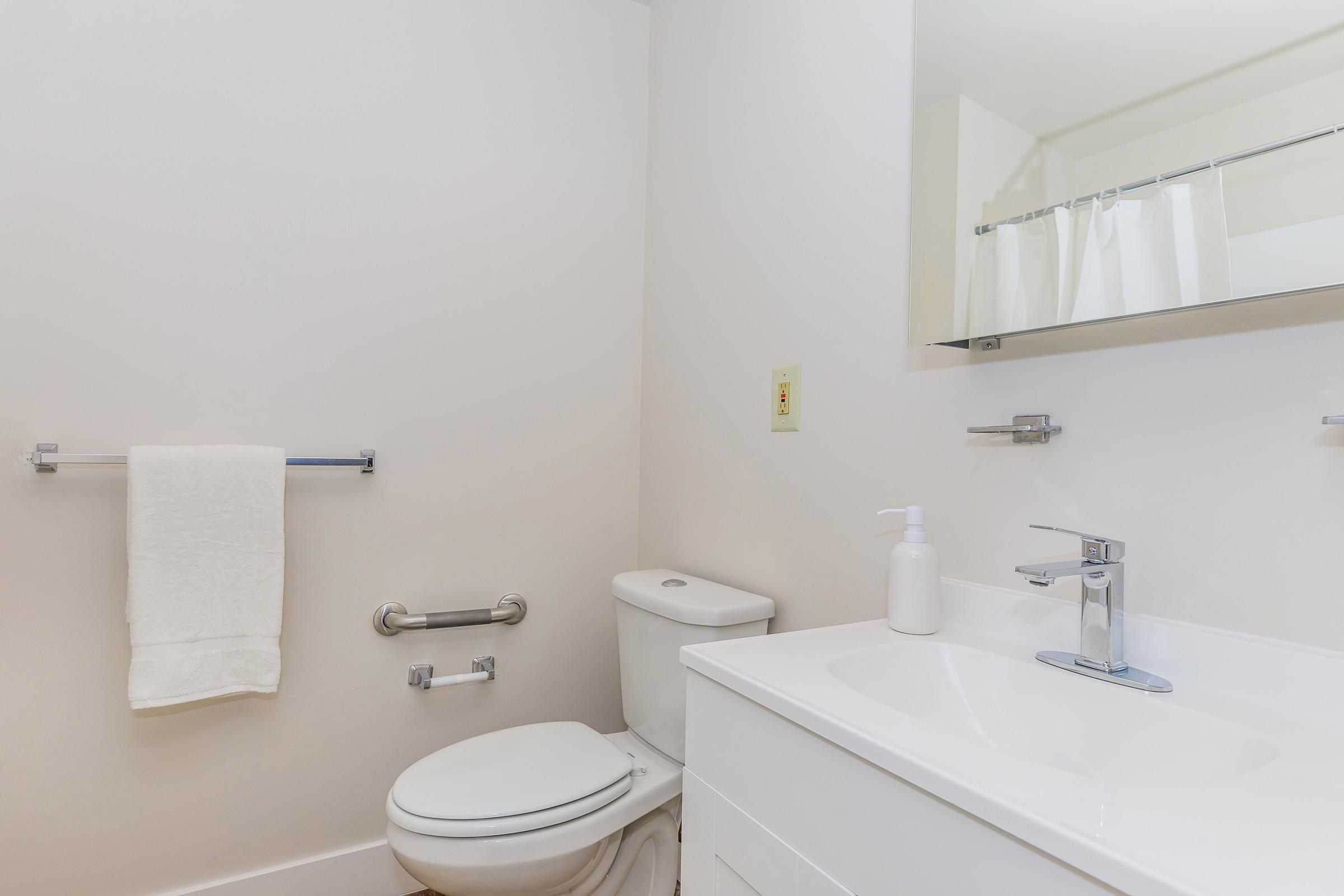 A clean, modern bathroom featuring a white toilet, sink with a sleek faucet, wall-mounted towel rack, and a mirror. The walls are painted light, and the space is well-lit, creating a fresh and minimalist atmosphere. A soap dispenser is placed next to the sink.