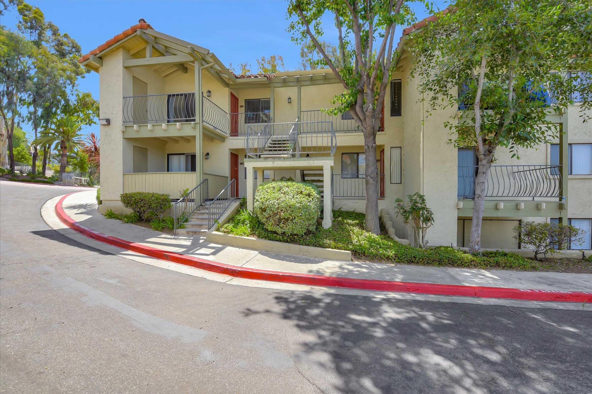 A well-maintained multi-unit apartment building with a gentle slope leading up to the entrance. Lush greenery surrounds the pathway, and the exterior features a light-colored facade with balconies. A red-curbed driveway adds a touch of color, and the atmosphere is bright and inviting under a clear blue sky.