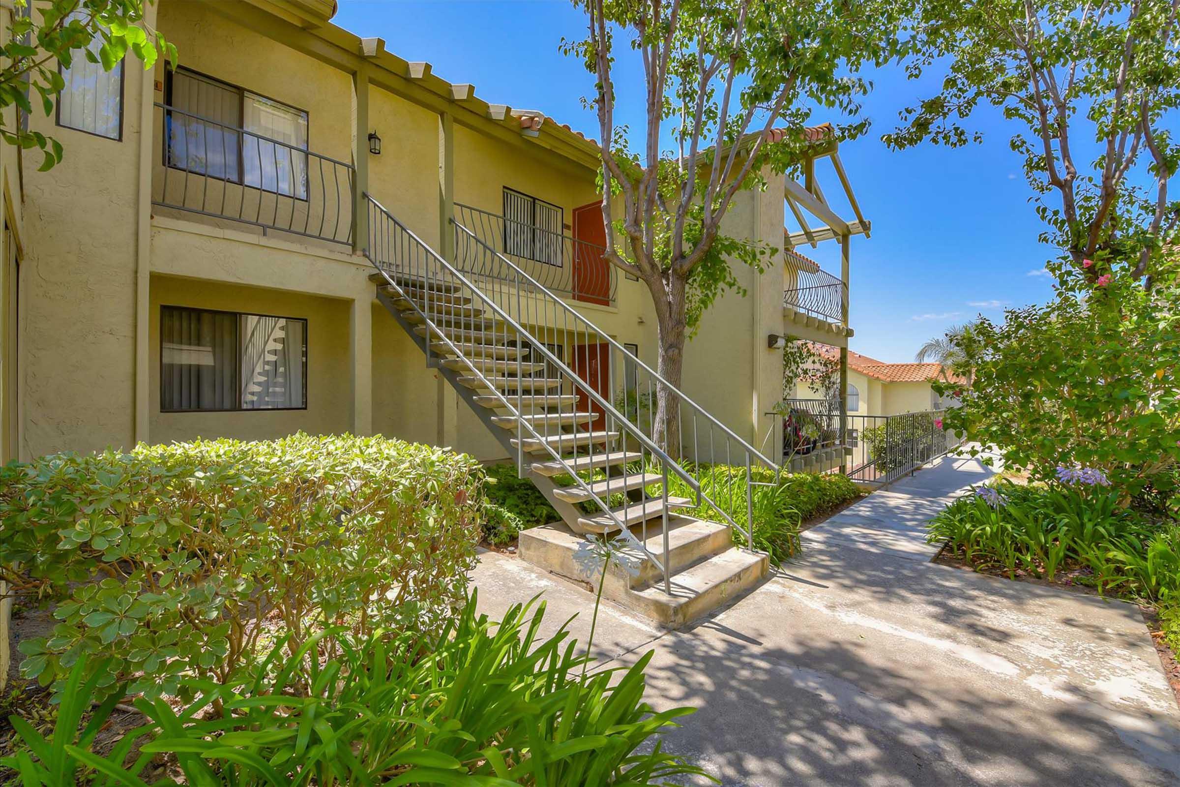 A view of a multi-unit apartment building featuring a set of metal stairs leading to the entrance. The structure is surrounded by green landscaping, including shrubs and small trees, under a clear blue sky. The pathway is paved and leads through well-maintained garden areas.