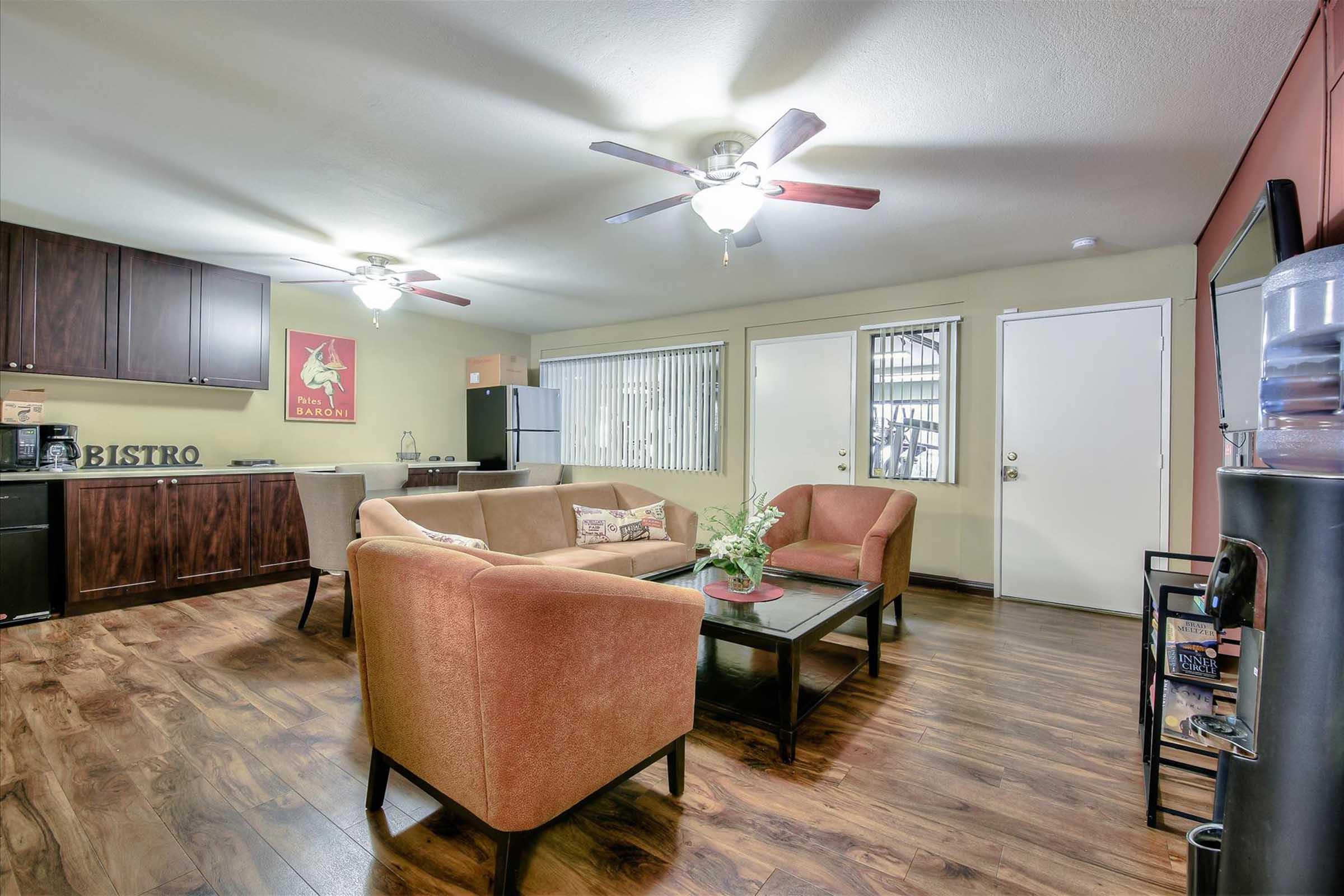 A cozy living room featuring two comfortable armchairs and a coffee table, with a small dining area in the background. Light-colored walls and wood flooring create a warm atmosphere. The space includes ceiling fans, large windows with blinds, and a kitchenette visible to one side.