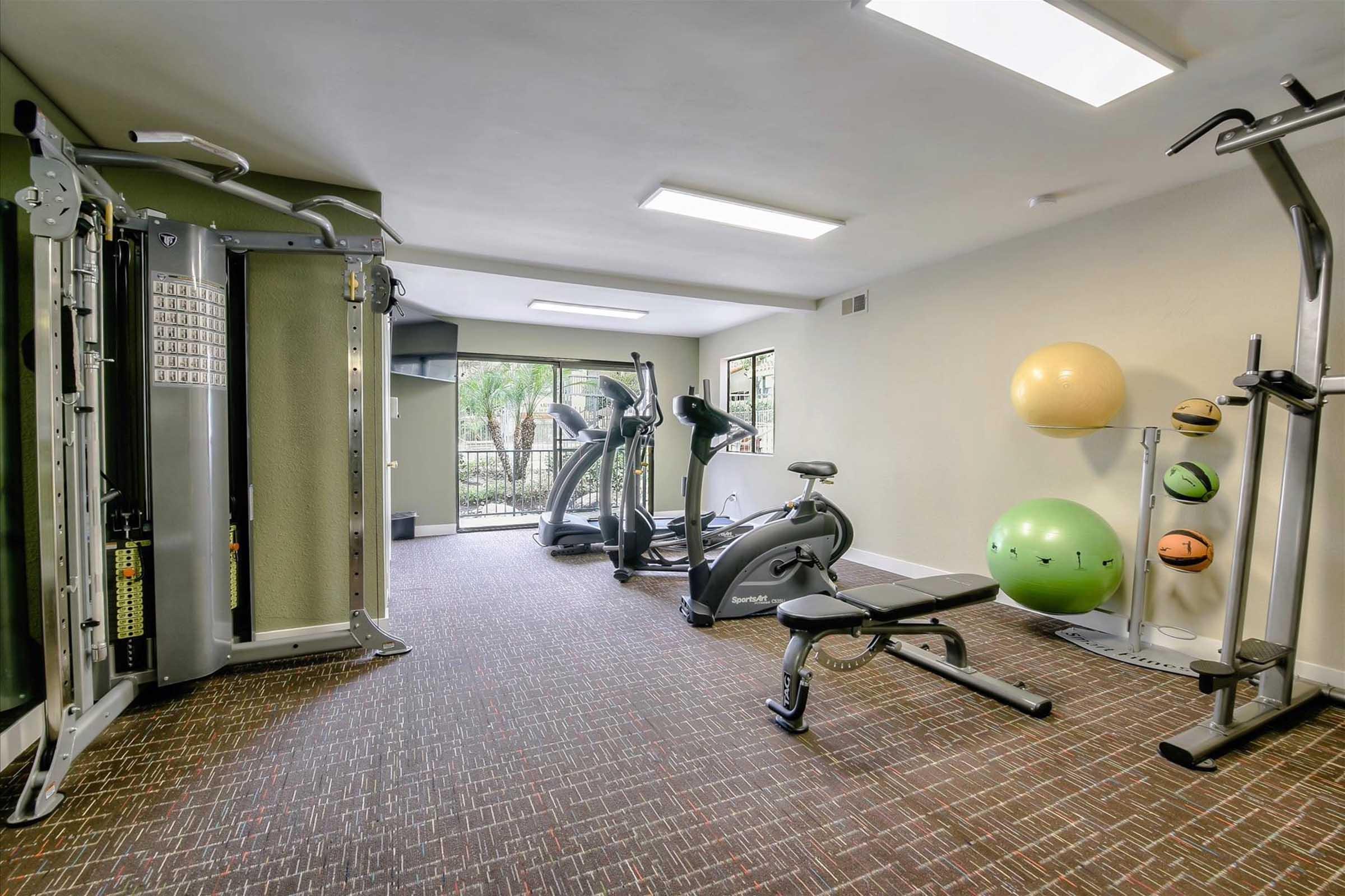 A modern gym interior featuring various exercise equipment, including a multi-gym station, elliptical machine, stationary bike, and a weight bench. Brightly lit with large windows, the space also includes fitness balls and wall-mounted storage for additional workout accessories.