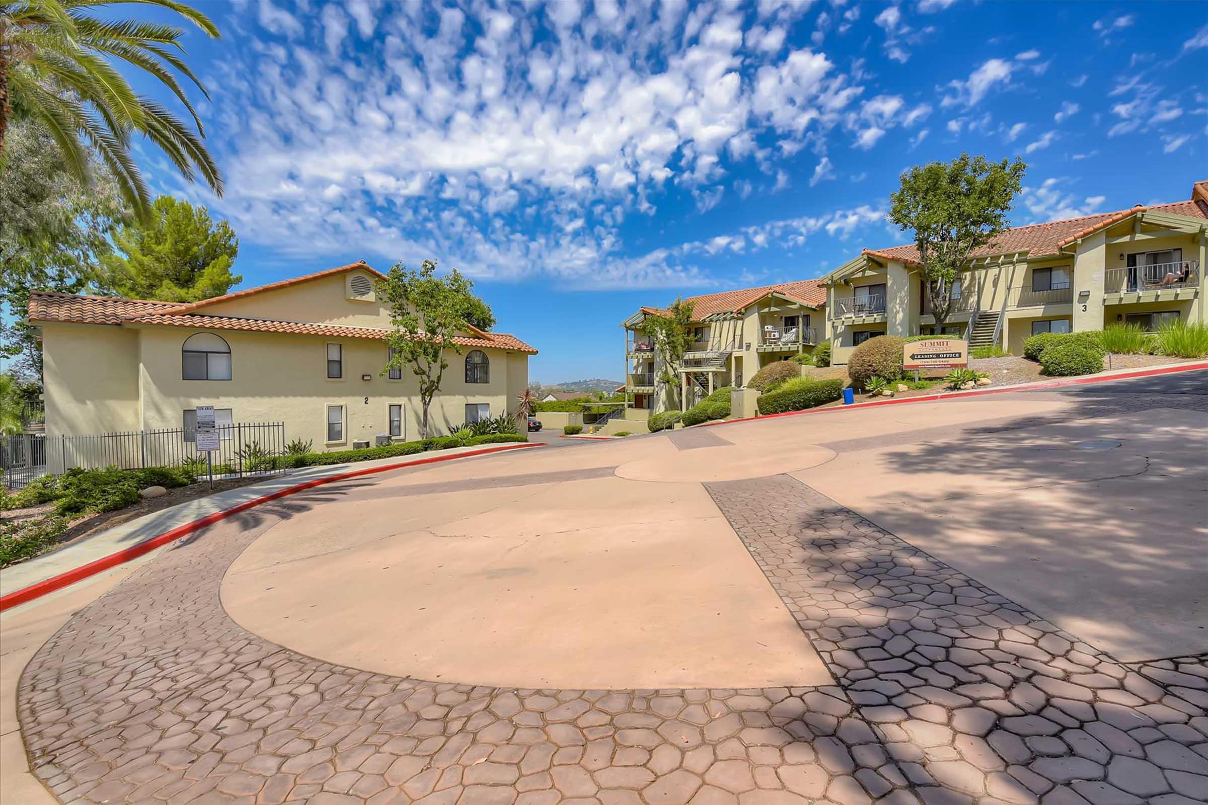 A view of a residential community with two apartment buildings on a curved driveway. Lush greenery and palm trees are present, and a bright blue sky with scattered clouds adds to the scenic atmosphere. The area appears well-maintained and inviting.