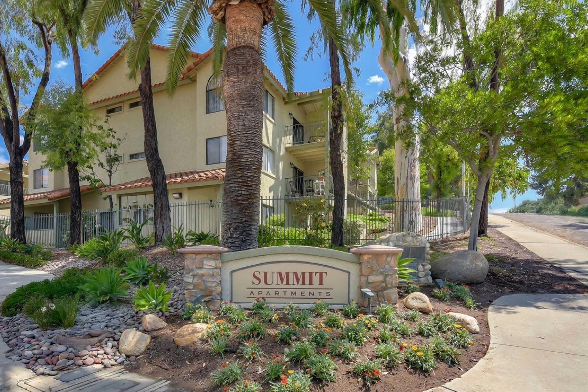 Sign for Summit Apartments in front of a multi-story building surrounded by palm trees and landscaped gardens. The walkway leading up to the entrance has decorative rocks and plants, with a clear blue sky in the background.