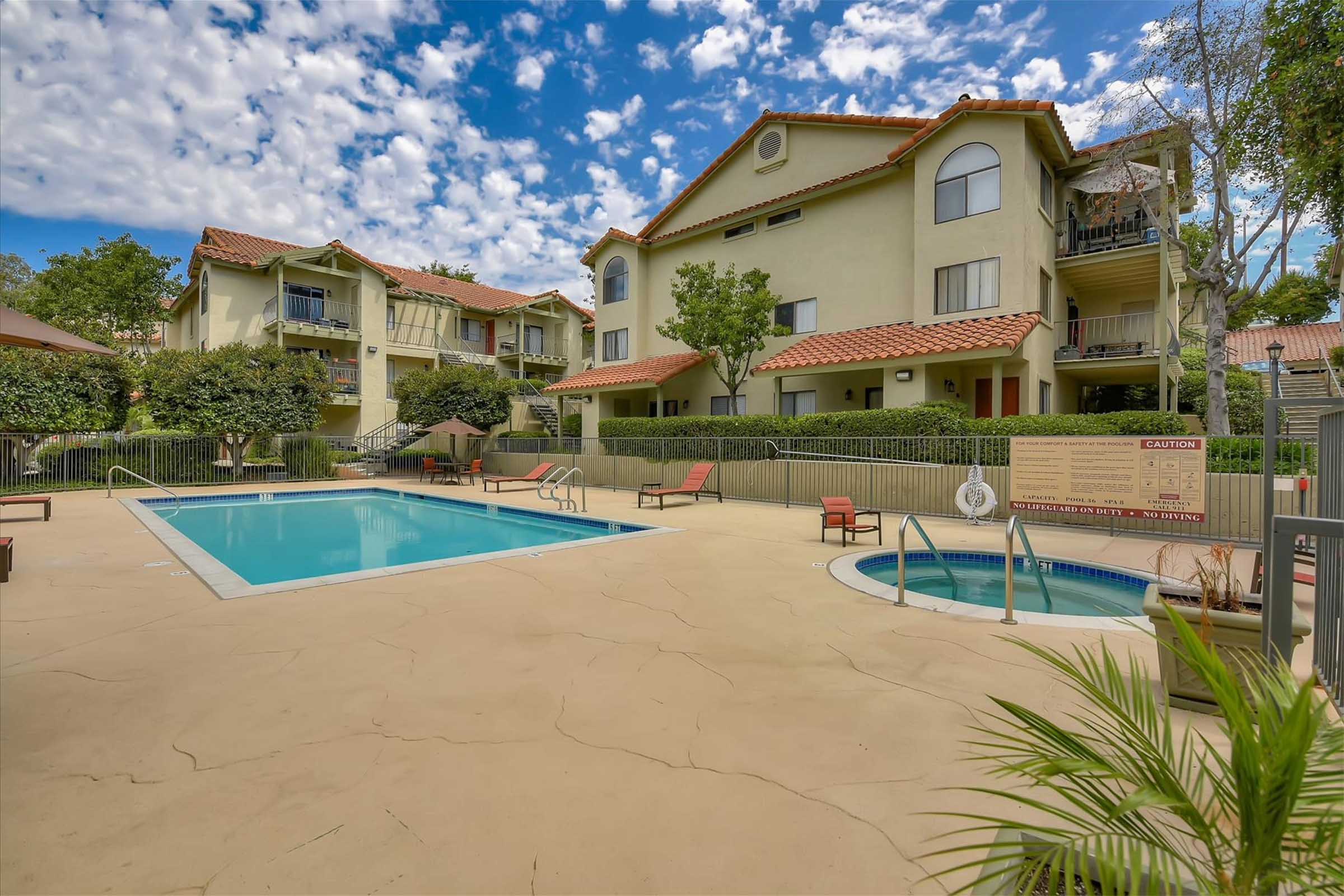 A clean swimming pool area surrounded by lounge chairs and a hot tub, with landscaped greenery and residential buildings in the background. The sky is partly cloudy, adding a serene atmosphere to the outdoor setting.